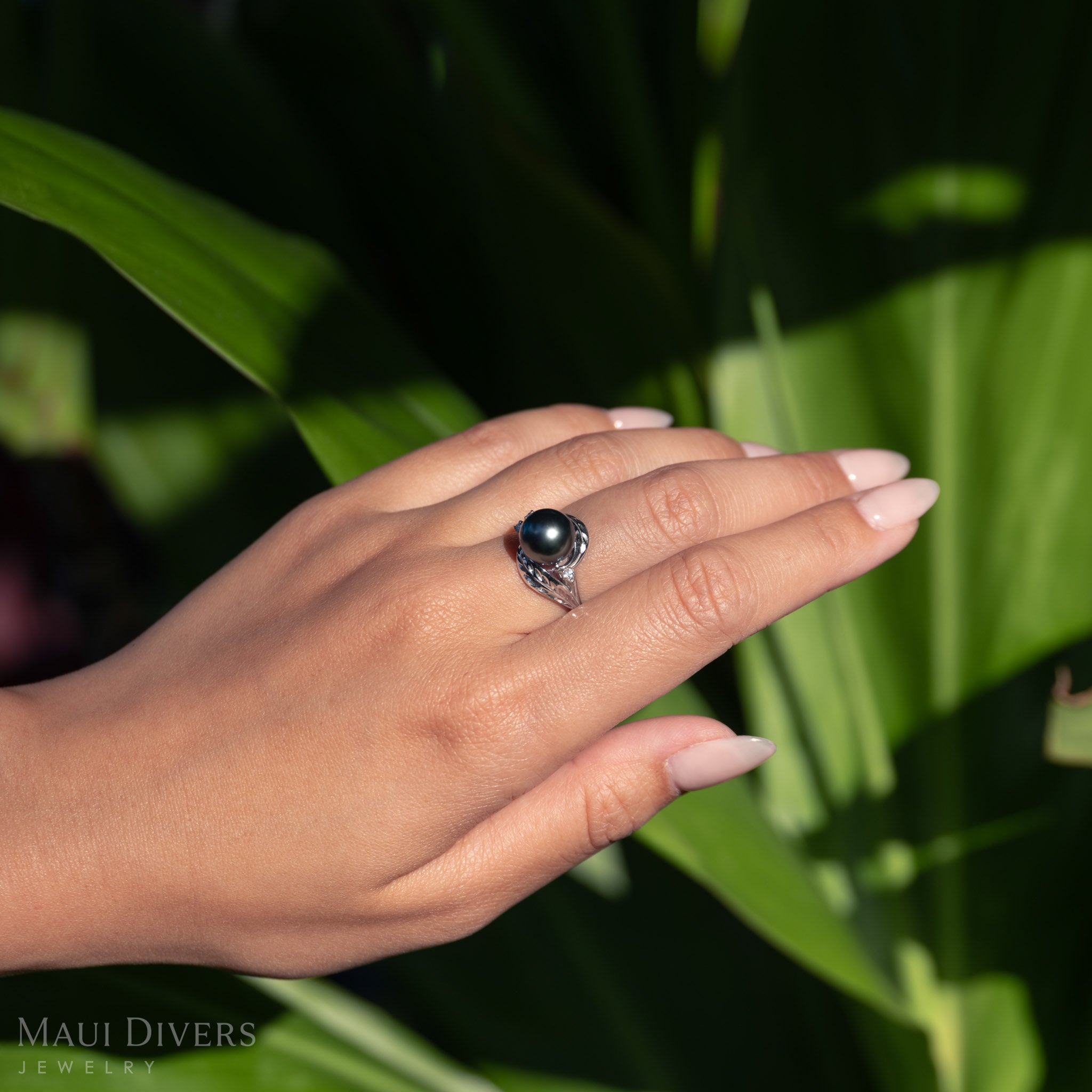 Hand wearing a Maile Leaf Tahitian black pearl ring against a green leafy background