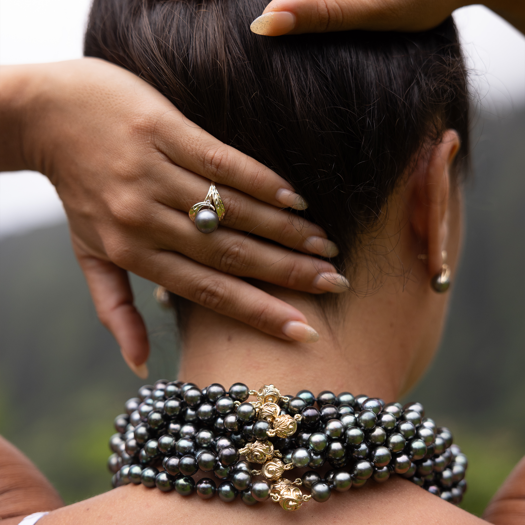 A woman's left hand touching the back of her neck with a maile leaf Tahitian black pearl ring in 14k yellow gold with diamonds.