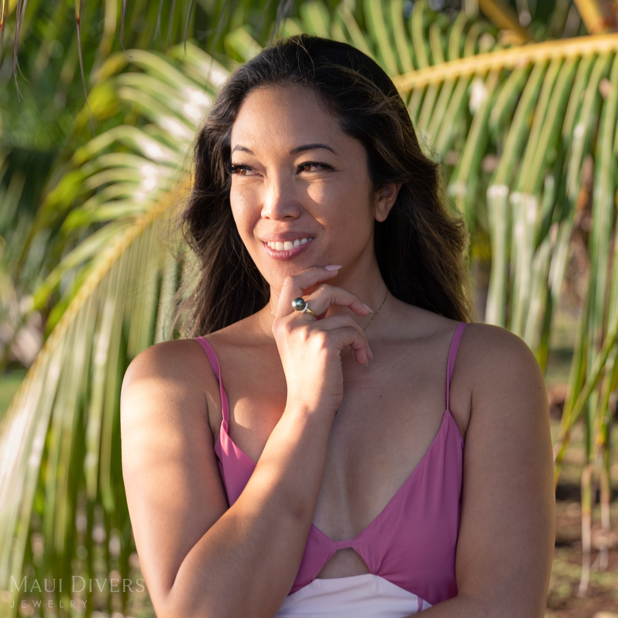Woman in a pink dress wearing a Tahitian black pearl ring with diamonds outdoors with palm trees in the background