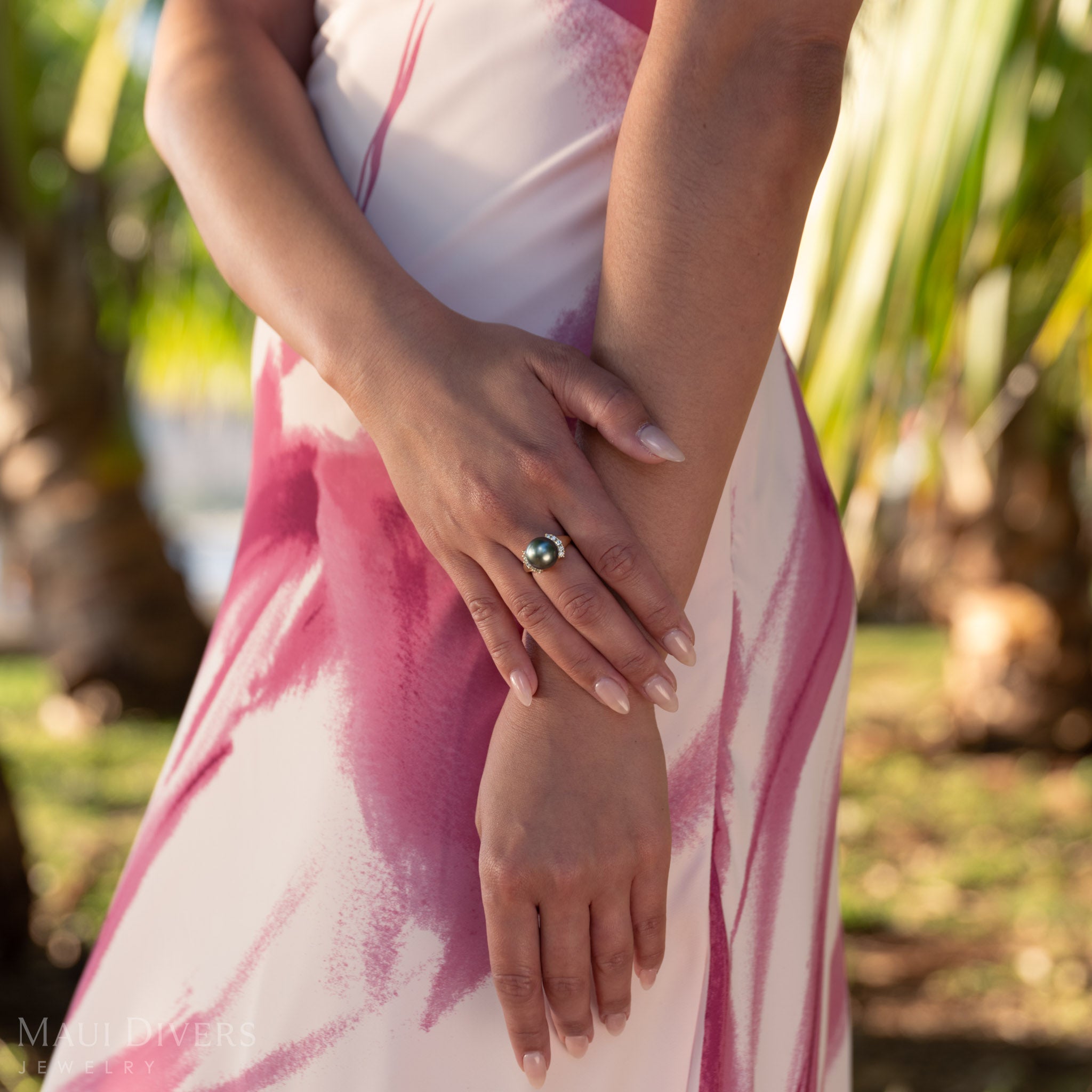 Person wearing a pink and white dress and a Tahitian black pearl ring in gold with diamonds, with a blurred natural background