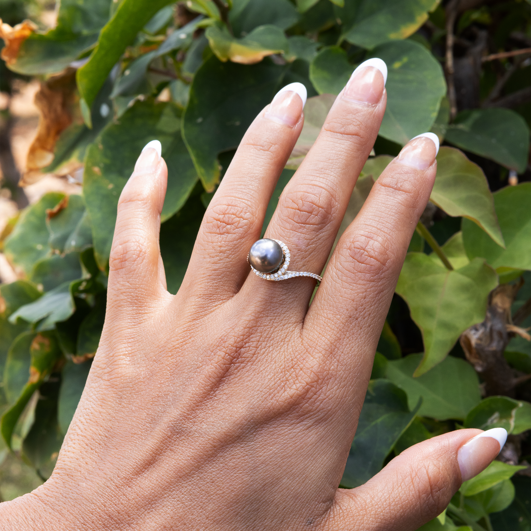 Close-up of a hand wearing a diamond halo, Tahitian black pearl ring over a leafy backdrop.