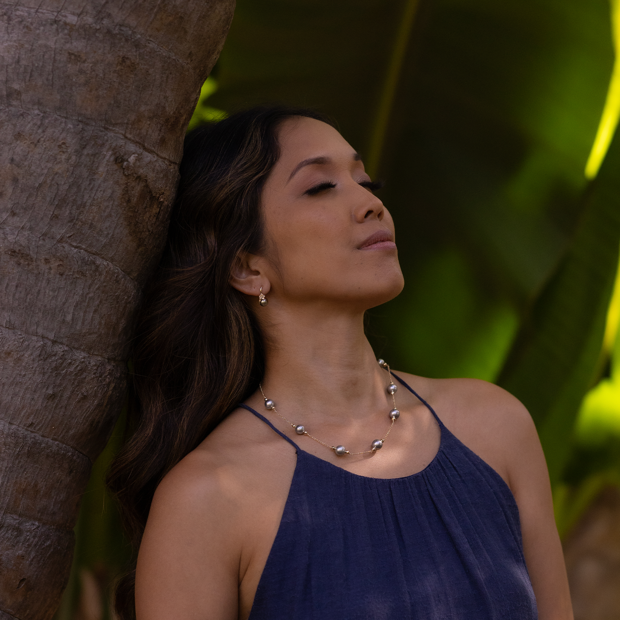 Woman leaning on a tree wearing a Tahitian black pearl necklace and plumeria earrings.