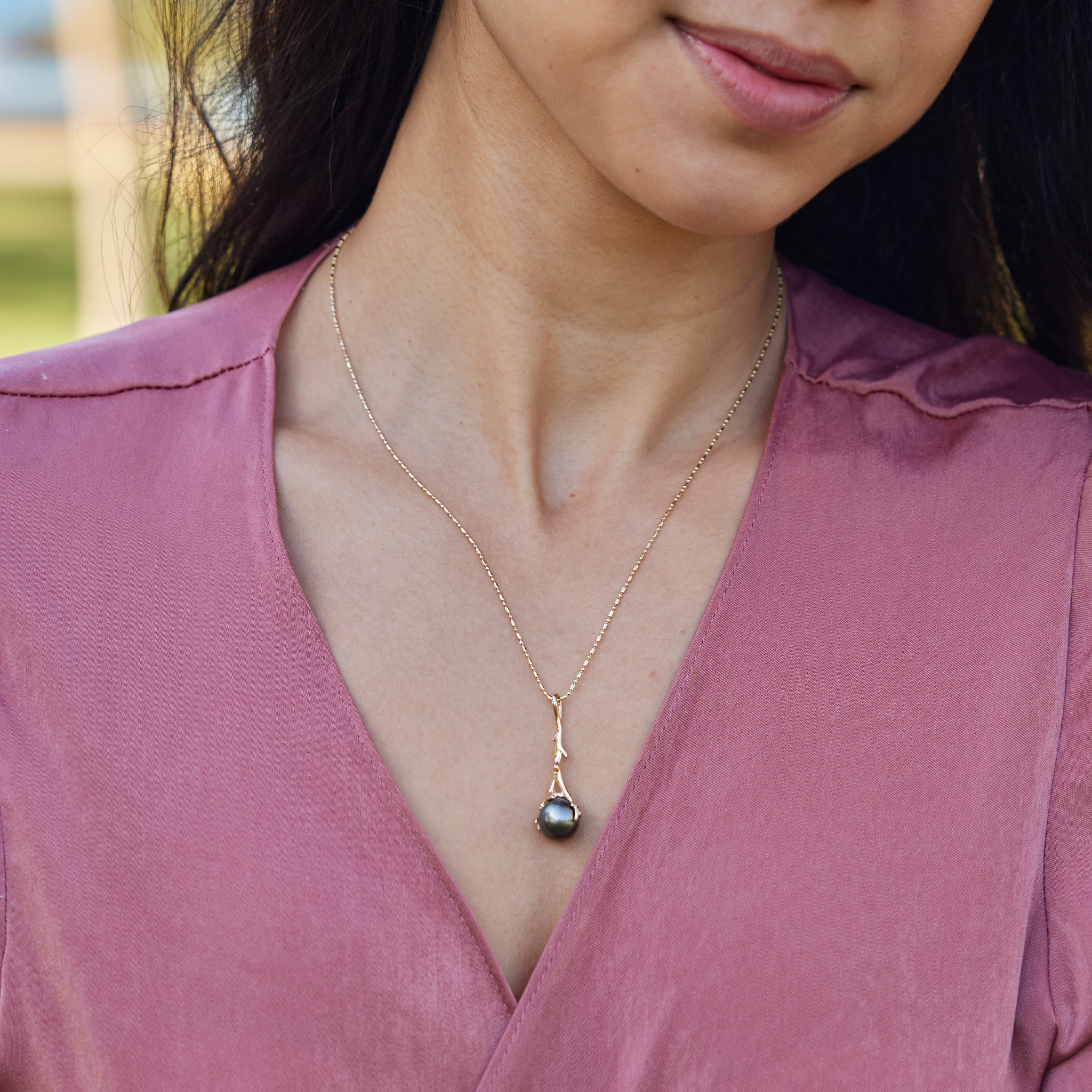 Person in a pink shirt wearing the Heritage Tahitian black pearl pendant in 14k yellow gold on a bar-ball chain