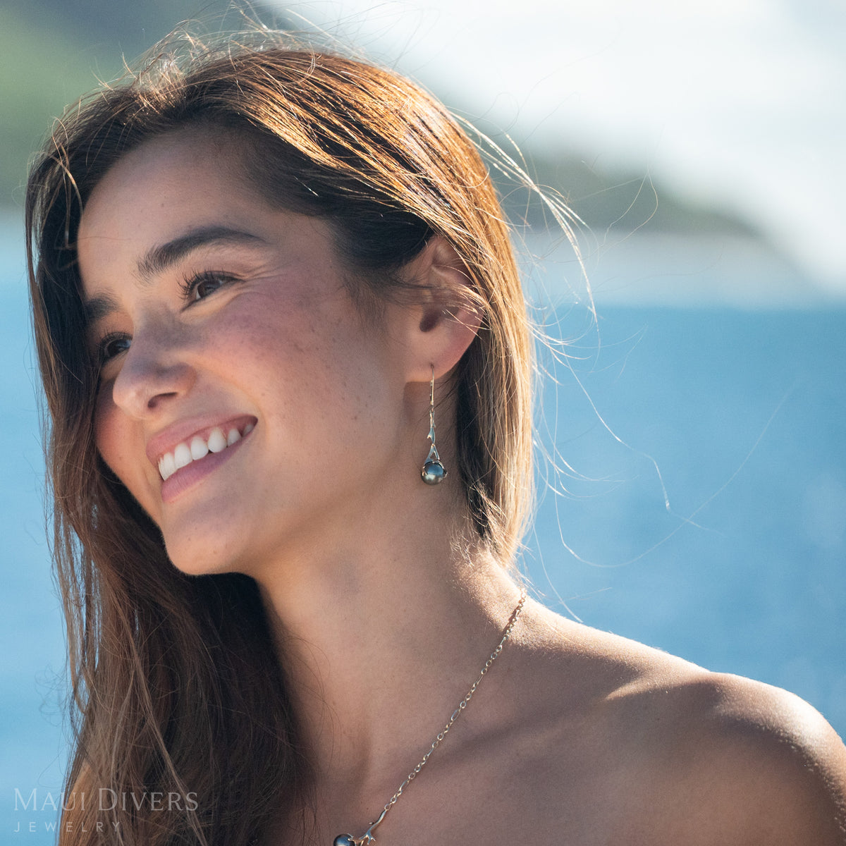 close-up of a woman wearing a gold Heritage Collection Leverback Earring with a Tahitian black pearl against a blurry blue mountain and ocean landscape