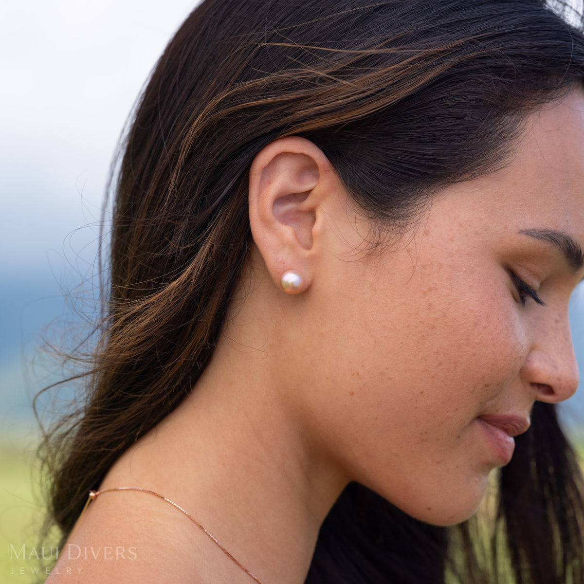 Close-up of a woman wearing a freshwater pearl natural color stud earring in 14k yellow gold with a blurry green and blue background