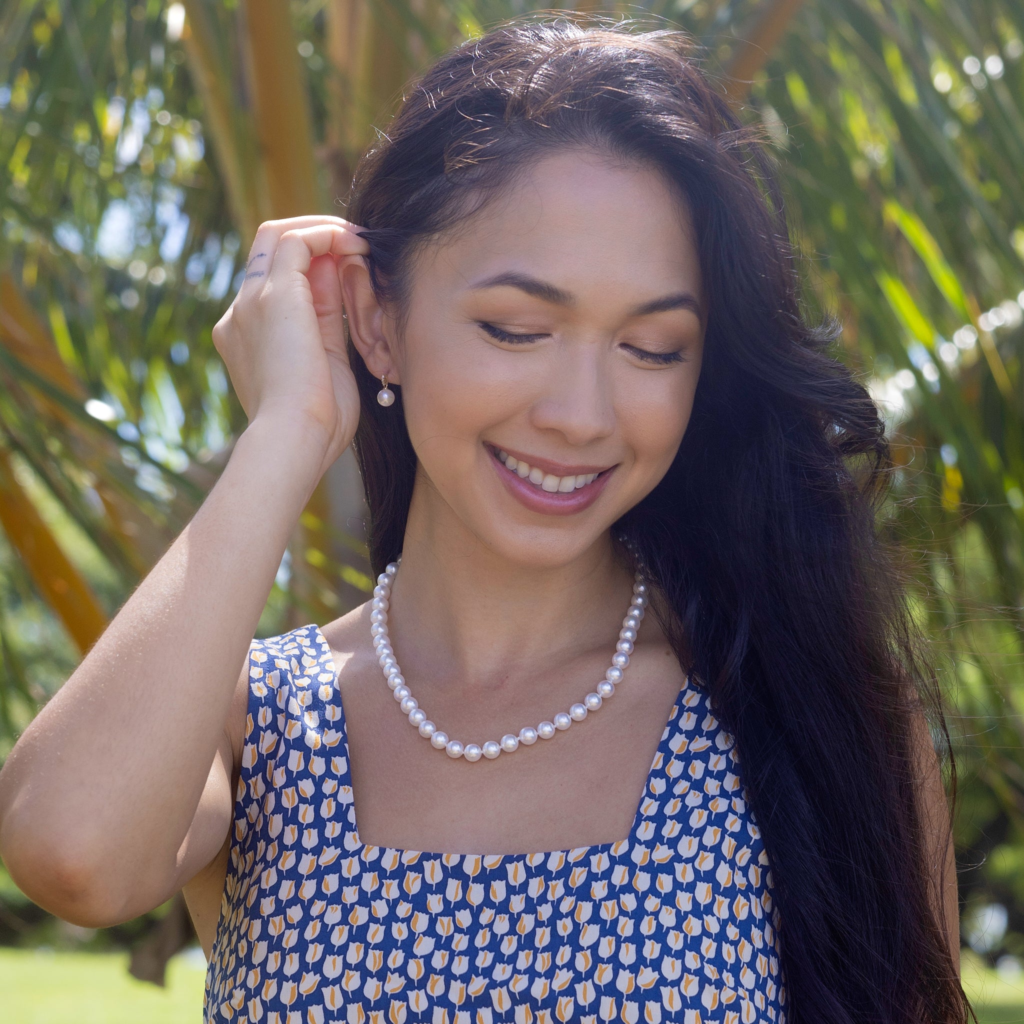 Smiling woman wearing an Akoya white pearl strand necklace and matching diamond-accented pearl earrings against a blurred background
