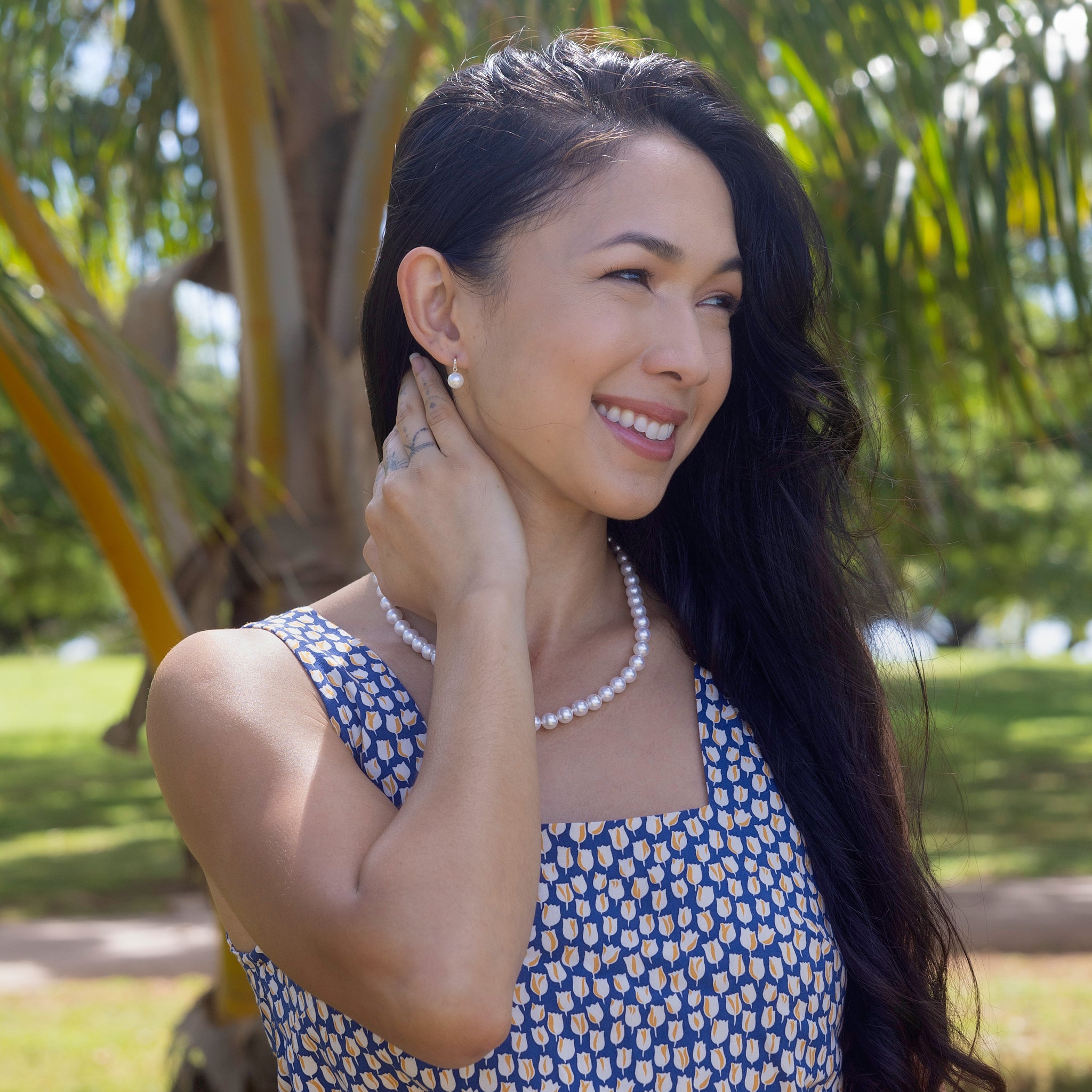Smiling woman wearing an Akoya white pearl strand necklace and matching diamond-accented pearl earrings against a blurred background