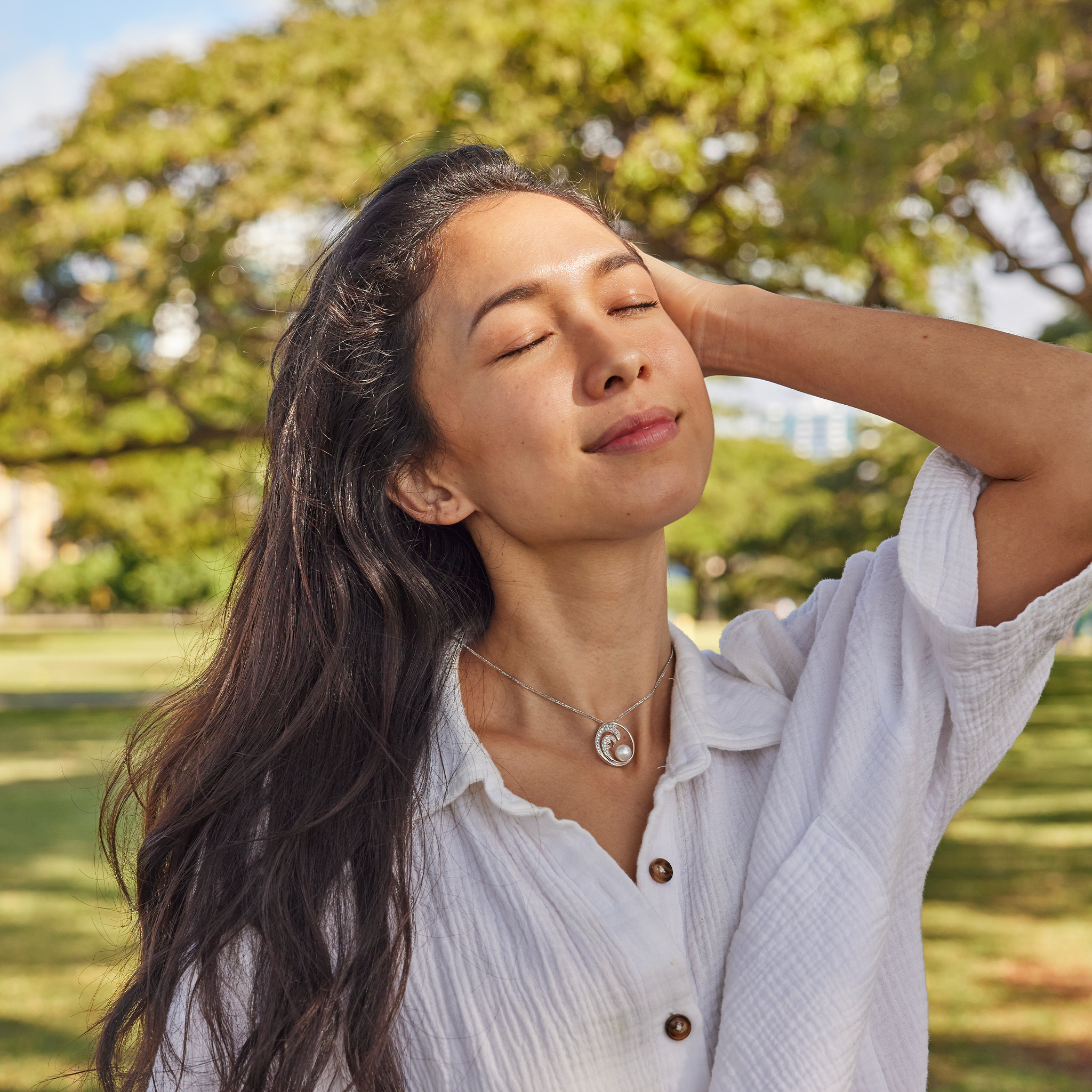 Smiling woman in a white shirt wearing a white gold diamond Nalu (Wave) pendant with an Akoya white pearl against a blurry grassy background with trees