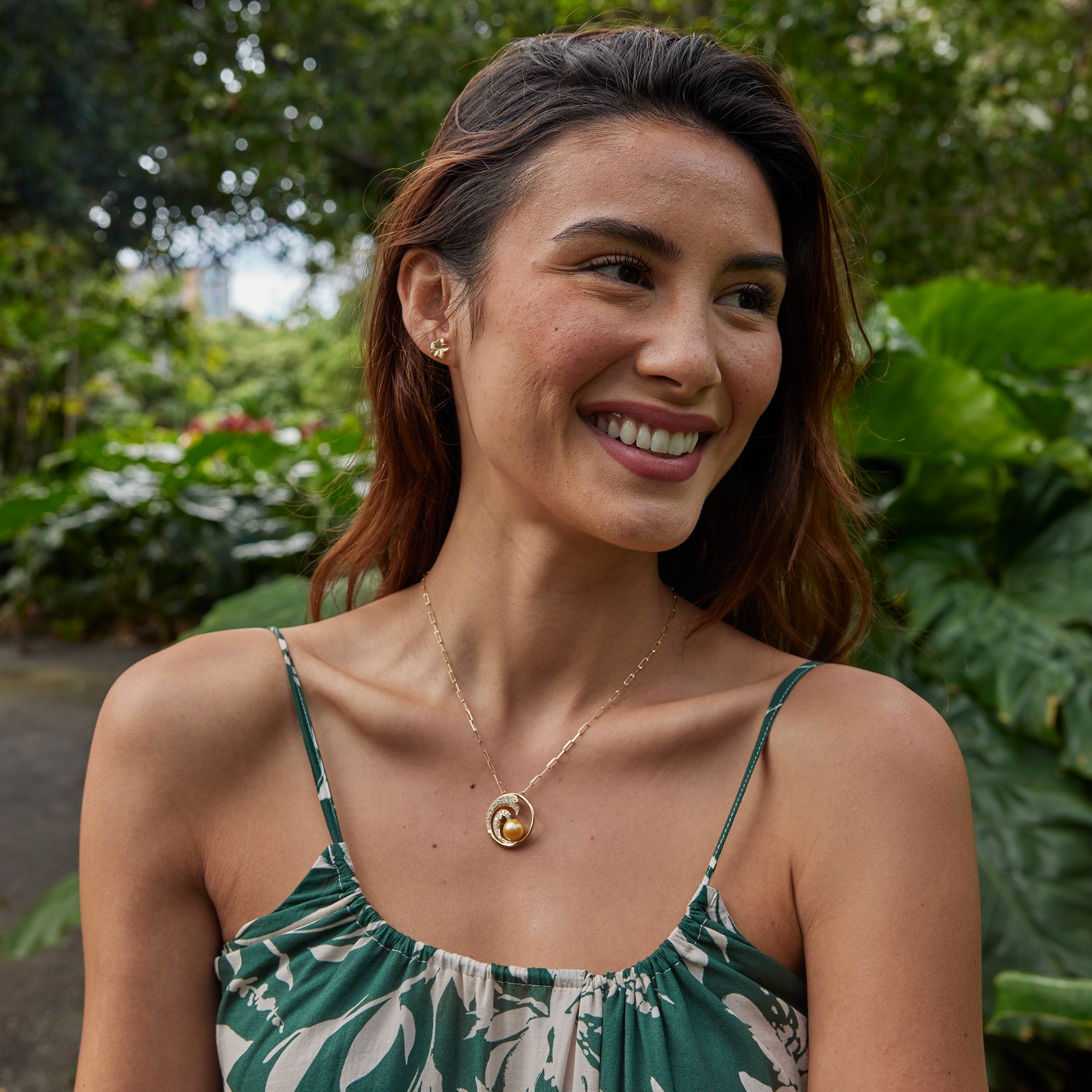 Smiling woman in a green patterned dress wearing gold plumeria earrings and a gold diamond wave South Sea gold pearl pendant on a paperclip chain and gold monstera earrings with a blurry garden in the background