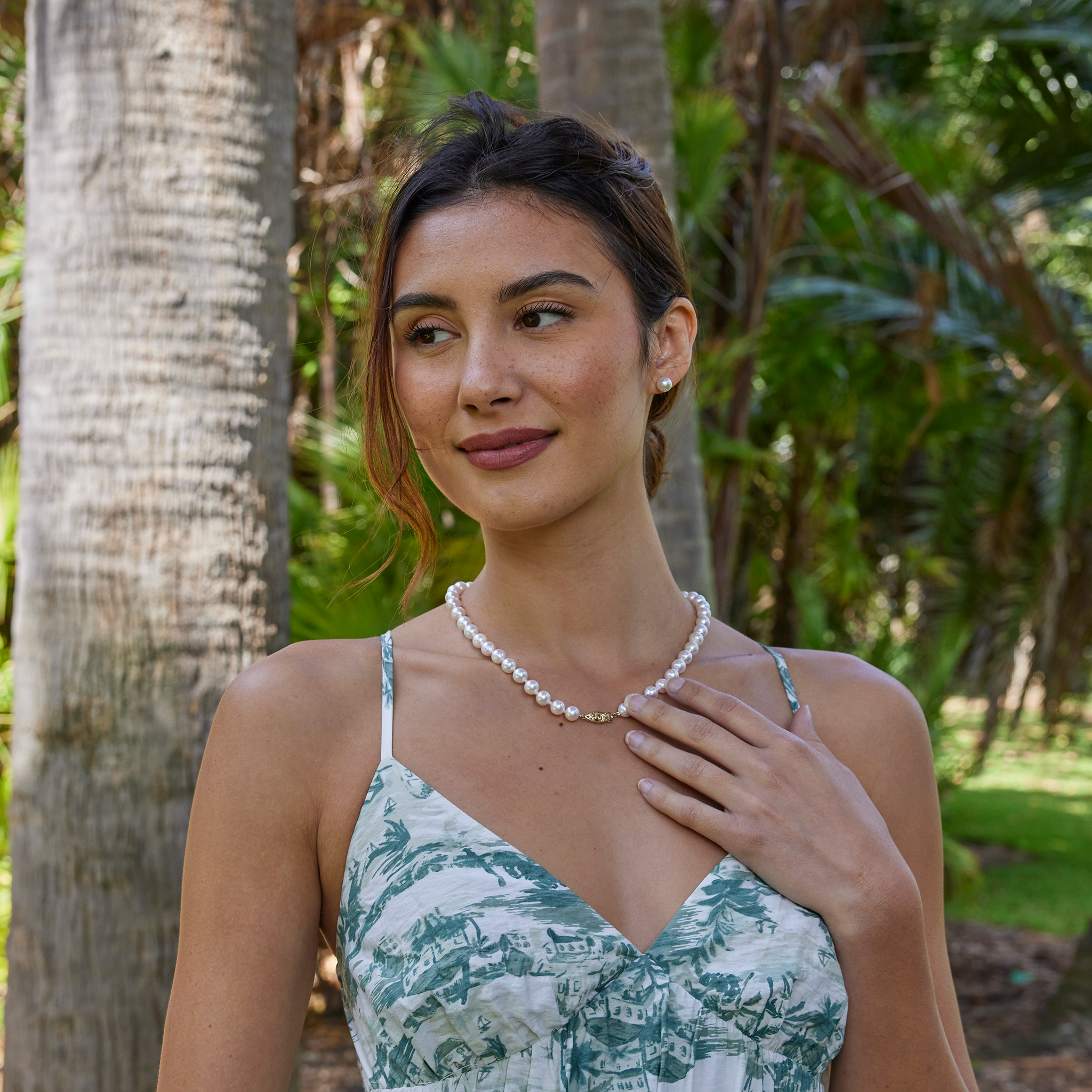 Smiling woman wearing an Akoya white pearl strand necklace with a gold clasp and matching pearl earrings against a blurred background