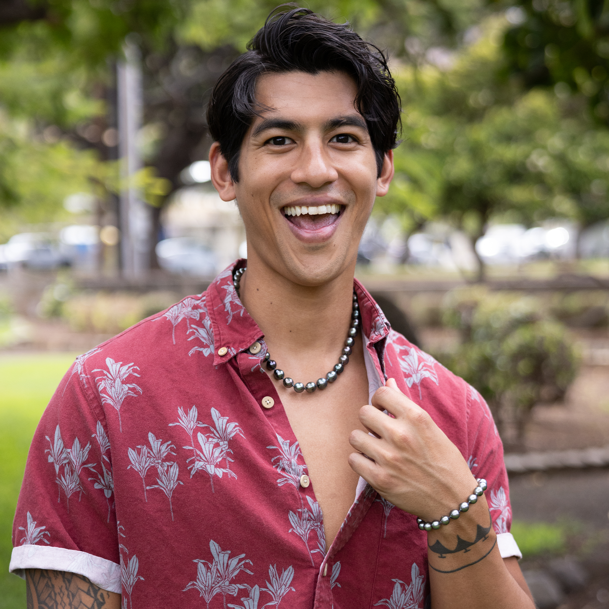 Smiling man in a red Aloha shirt wearing a Tahitian black pearl strand necklace and matching single-strand bracelet against a blurred background of trees