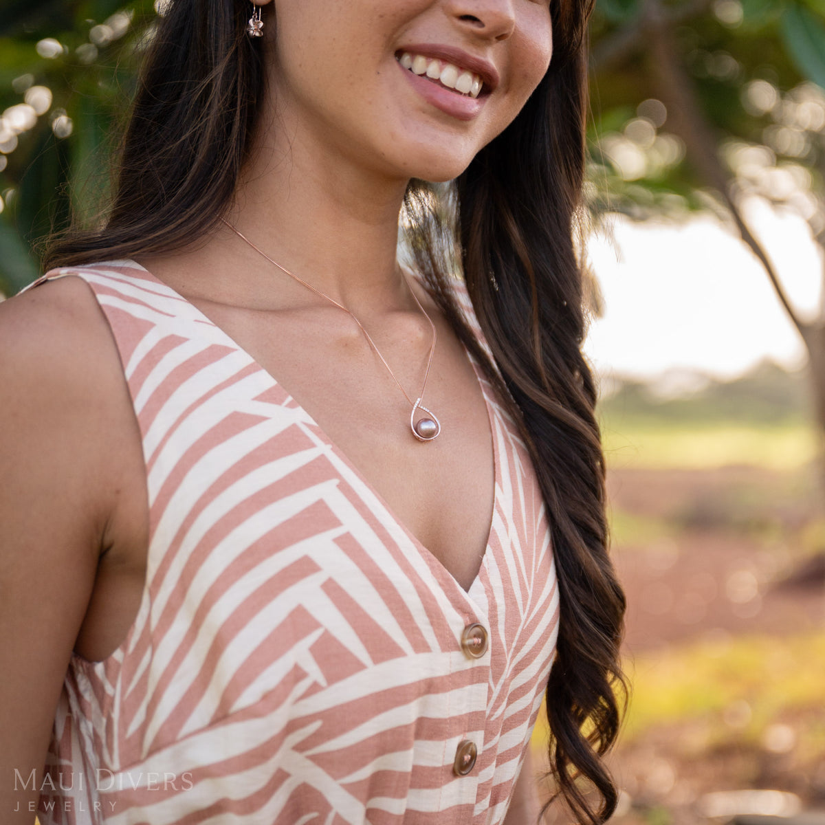 A smiling woman wearing a teardrop-shaped Lilac Freshwater Pearl Pendant in 14k Rose Gold with a pink and white top. She is turned on an angle, and blurred green trees and green grass are seen in the background.