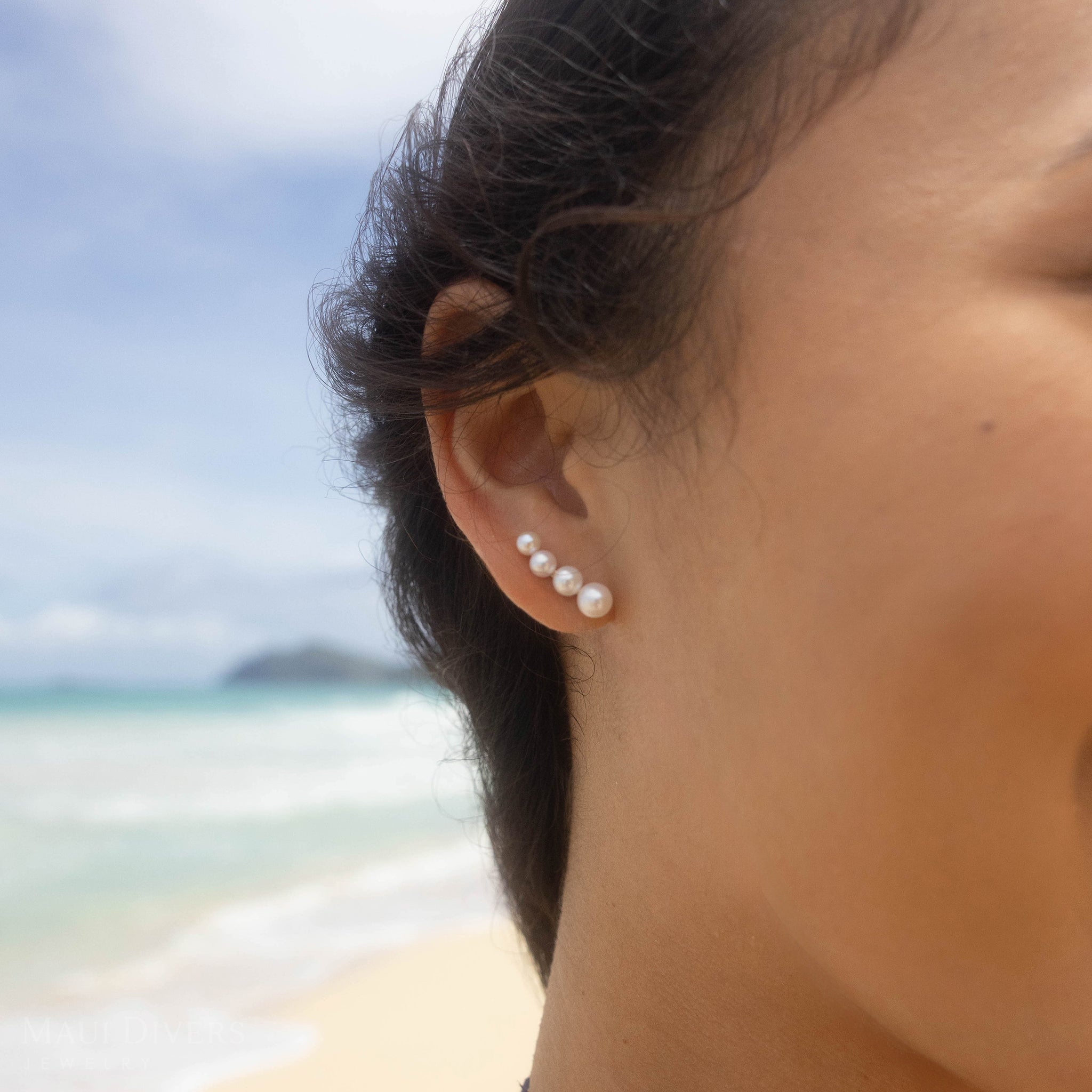 Close-up of a four-pearl gradient earring with Akoya and freshwater white pearls worn on an ear against a blurred beach background