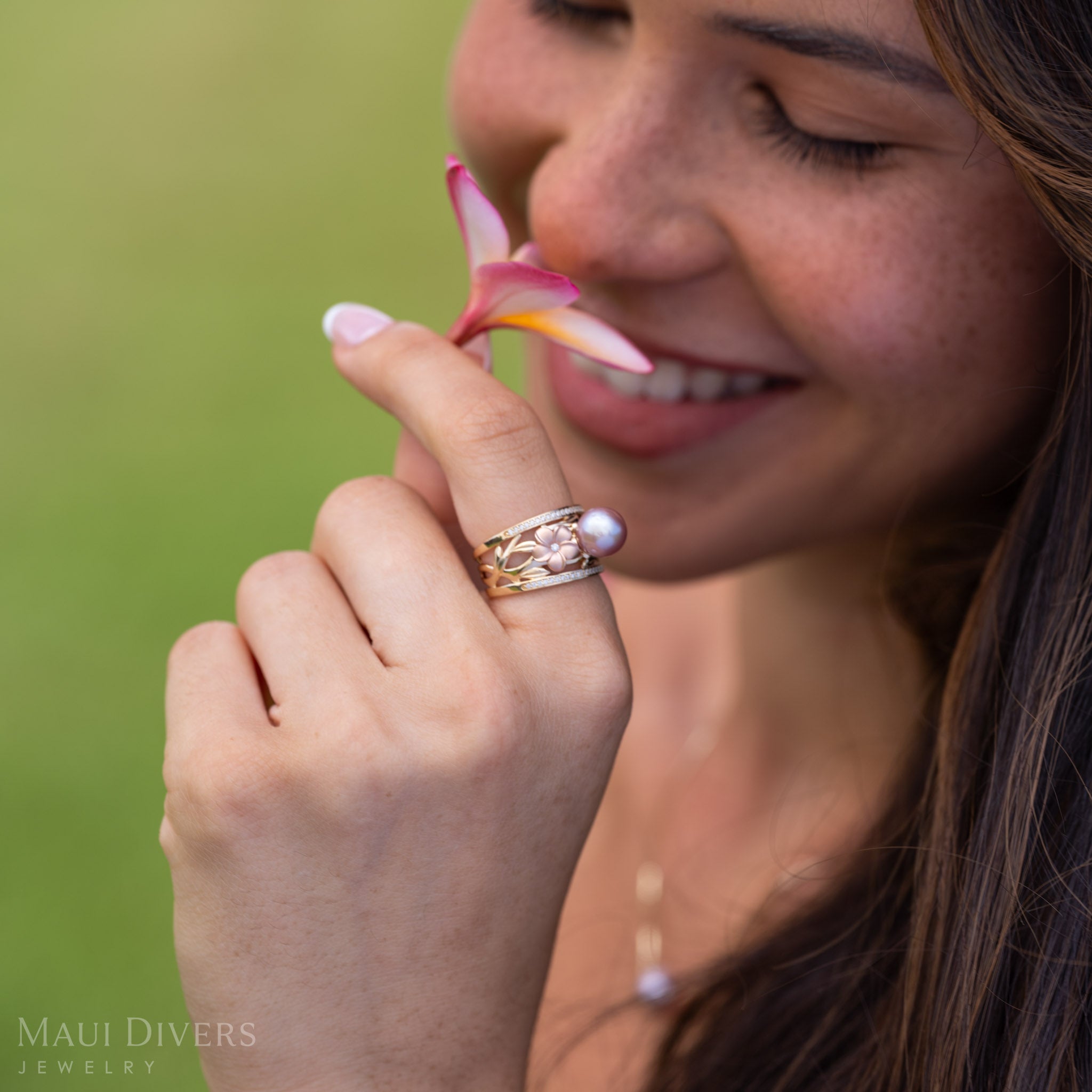 A smiling woman wearing a Natural Lavender Freshwater Pearl Ring with Tri Color Plumerias in 14k Gold accented with Diamonds smelling a light pink and yellow plumeria flower.