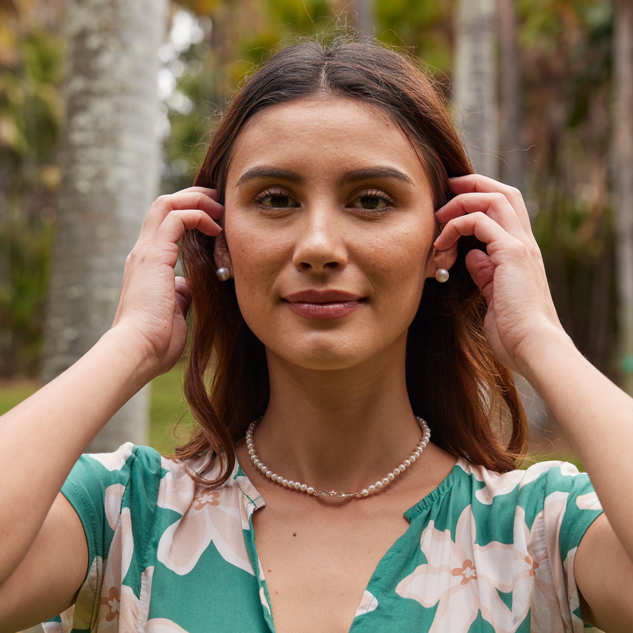Woman in a floral-patterned dress wearing white pearl stud earrings and the Hawaiian Heirloom Plumeria Freshwater White Pearl Necklace as she adjusts her hair