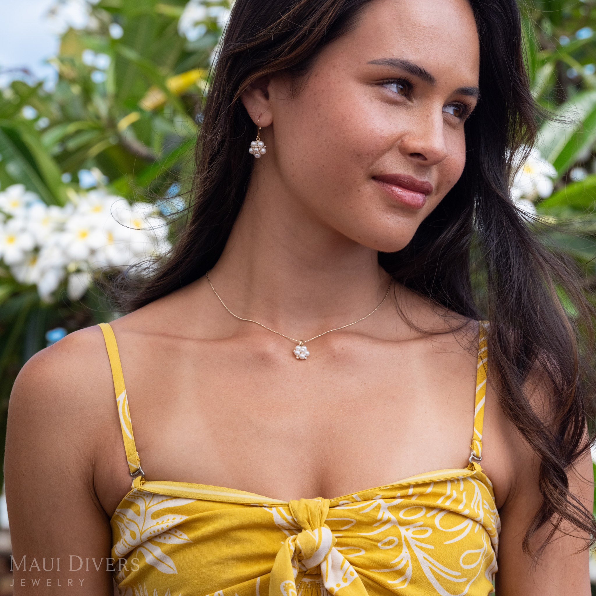 Side-profile of a woman wearing Tiny Natural Freshwater White Pearls on a 14k Gold earring accented with Diamonds, in the background there are green and white plumeria trees.