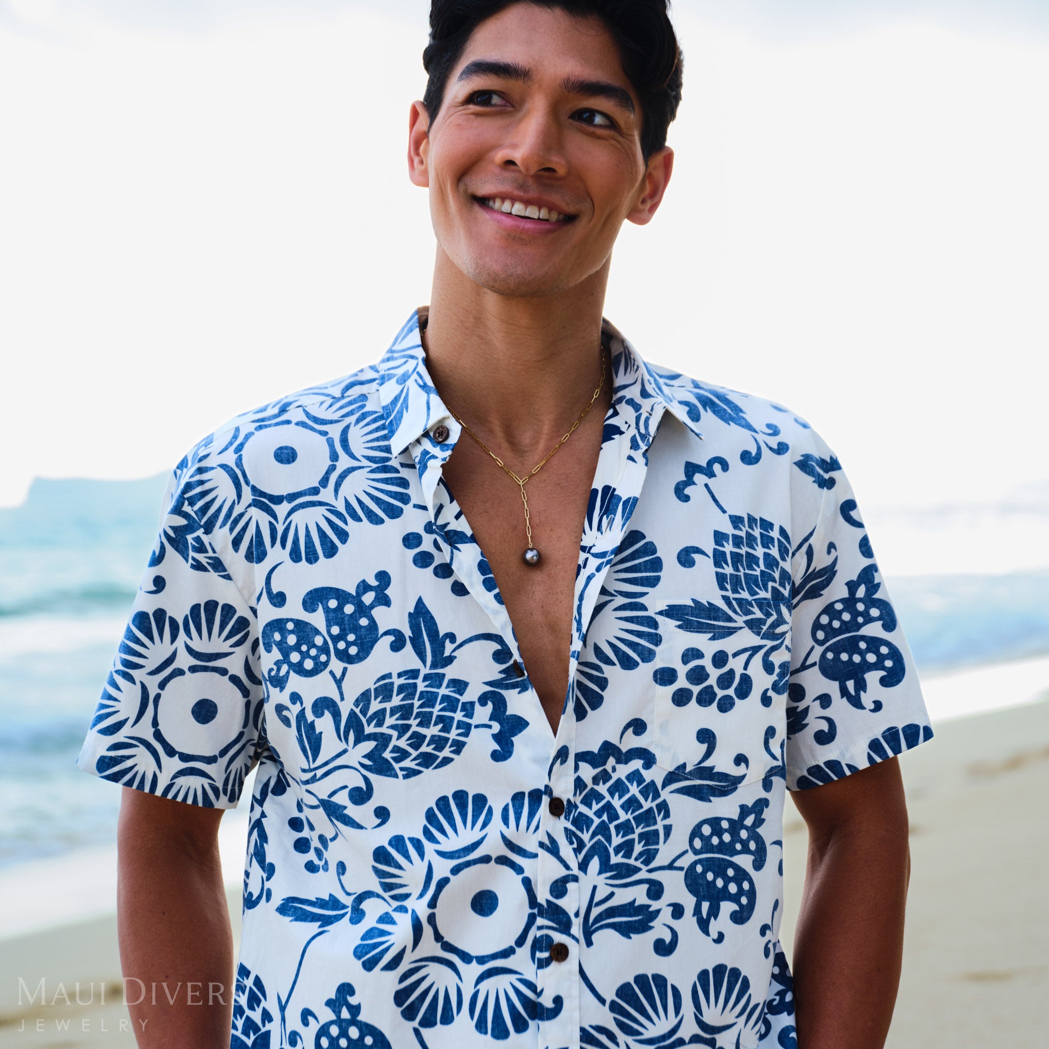 Man wearing a blue and white patterned shirt and a Tahitian black pearl necklace on a beach