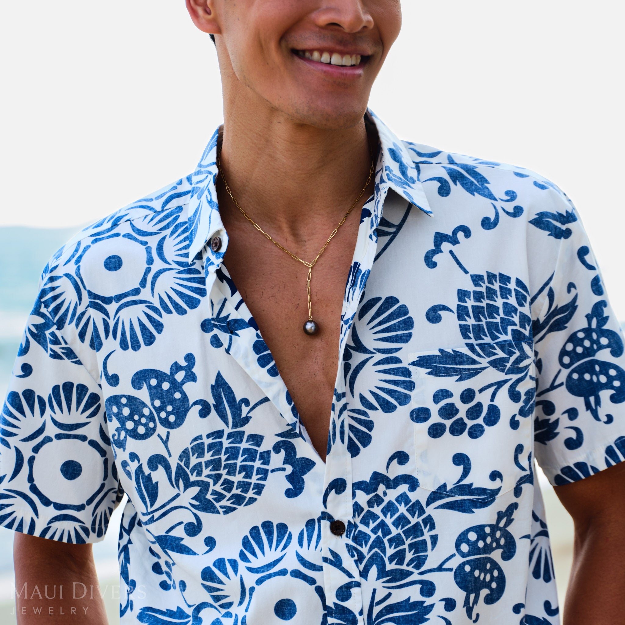 Man wearing a blue and white patterned shirt with a Tahitian black pearl necklace, standing outdoors.