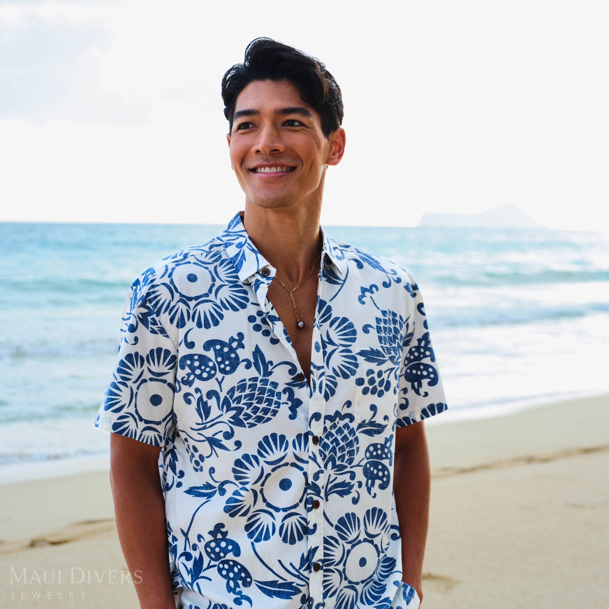Man wearing a blue and white patterned shirt and a Tahitian black pearl necklace on a beach