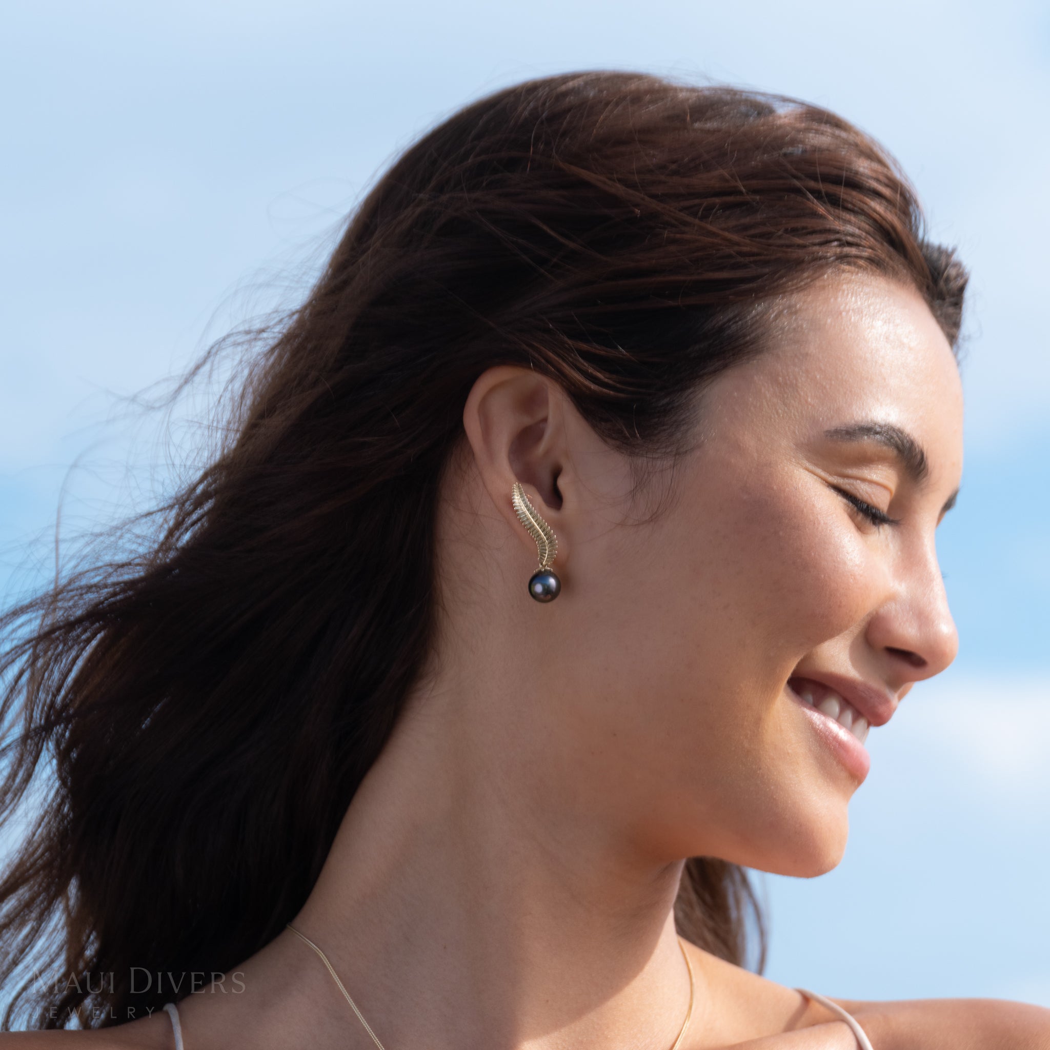 Close-up of a smiling woman wearing the 14k yellow gold Kupukupu fern Tahitian black pearl climber style earrings with a blue sky background