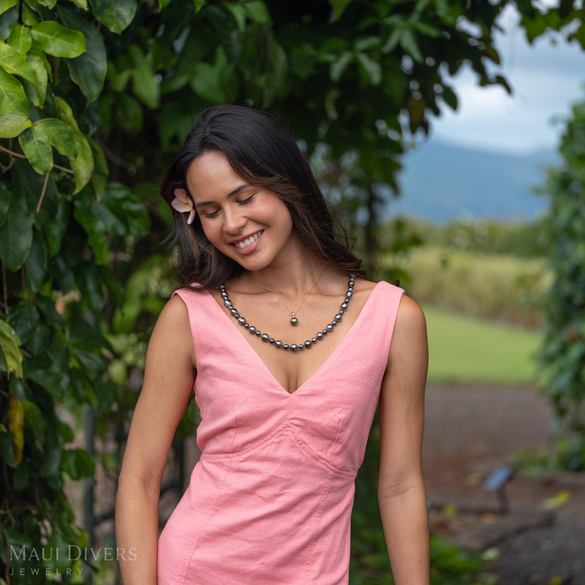 A woman smiling wearing a Natural Tahitian Black Pearl Strand Necklace paired with a single Tahitian Black Pearl Necklace. She is in a pink dress against a green leafy background.