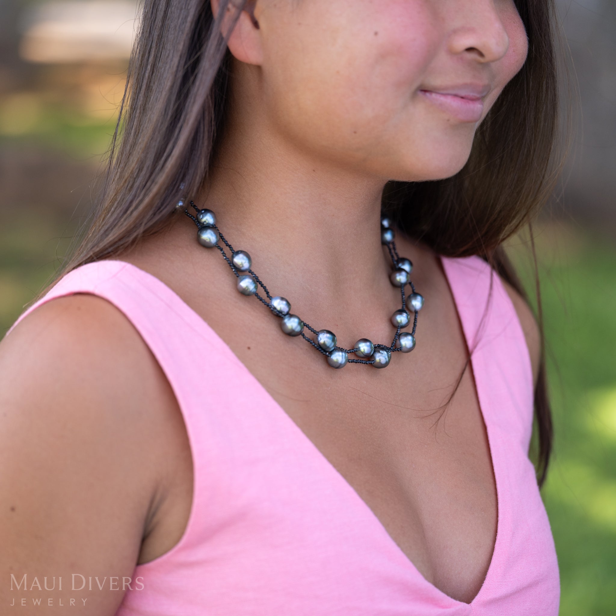A close-up of a smiling woman in a light pink top wearing a Tahitian Black Pearl and Black Spinel Layered Necklace with a 14k yellow gold clasp, against a blurred green grass background