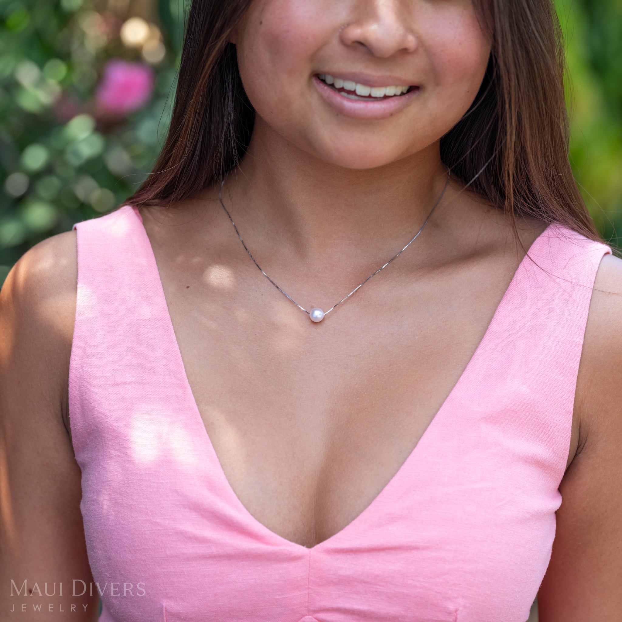 A close-up sideview of a woman in a light pink top wearing an Akoya White Pearl Necklace in 14k white gold against a blurred hibiscus plant background