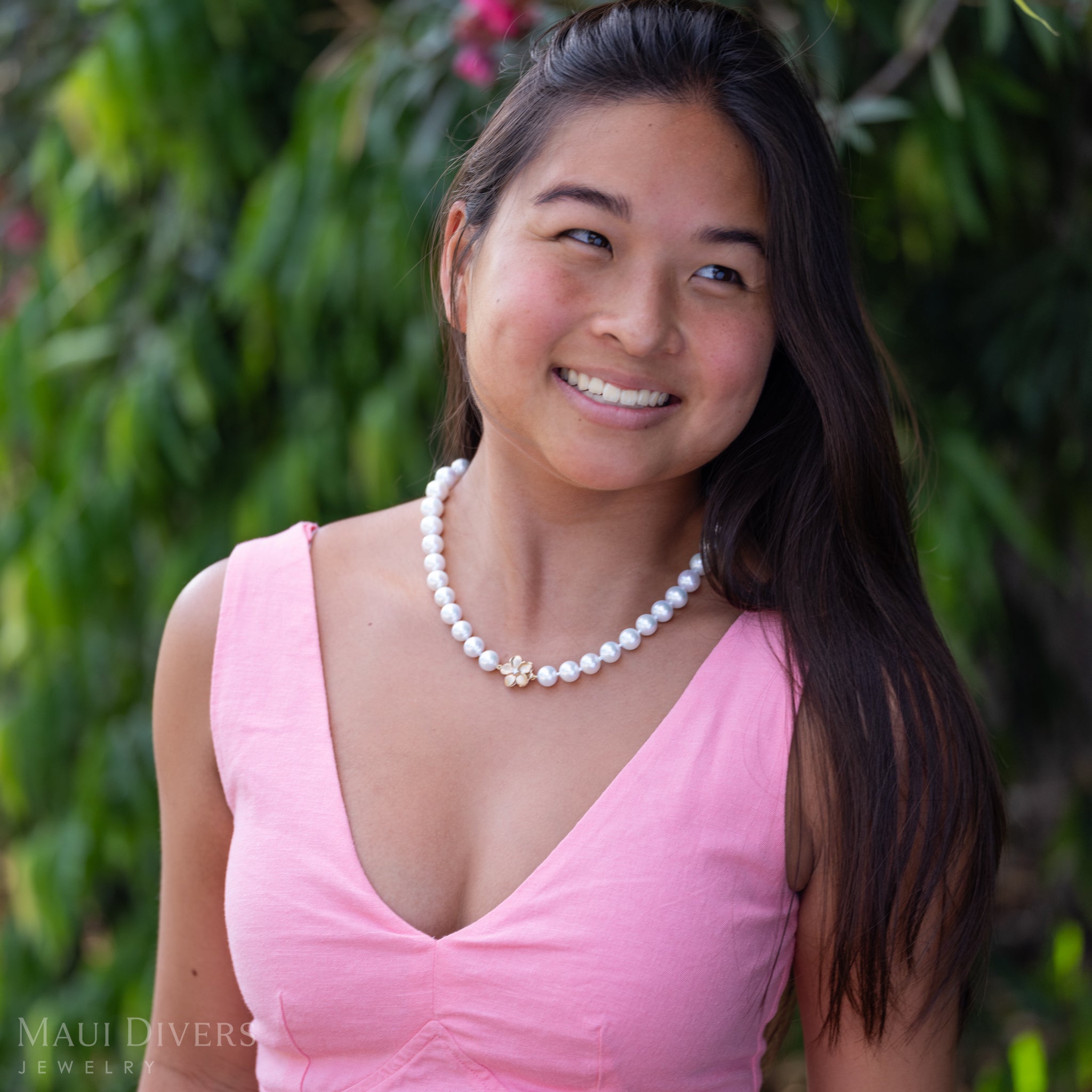 A smiling woman in a light pink top wearing a Freshwater White Pearl Strand with Diamond Plumeria Clasp in 14k Yellow Gold, against a blurred leafy green background