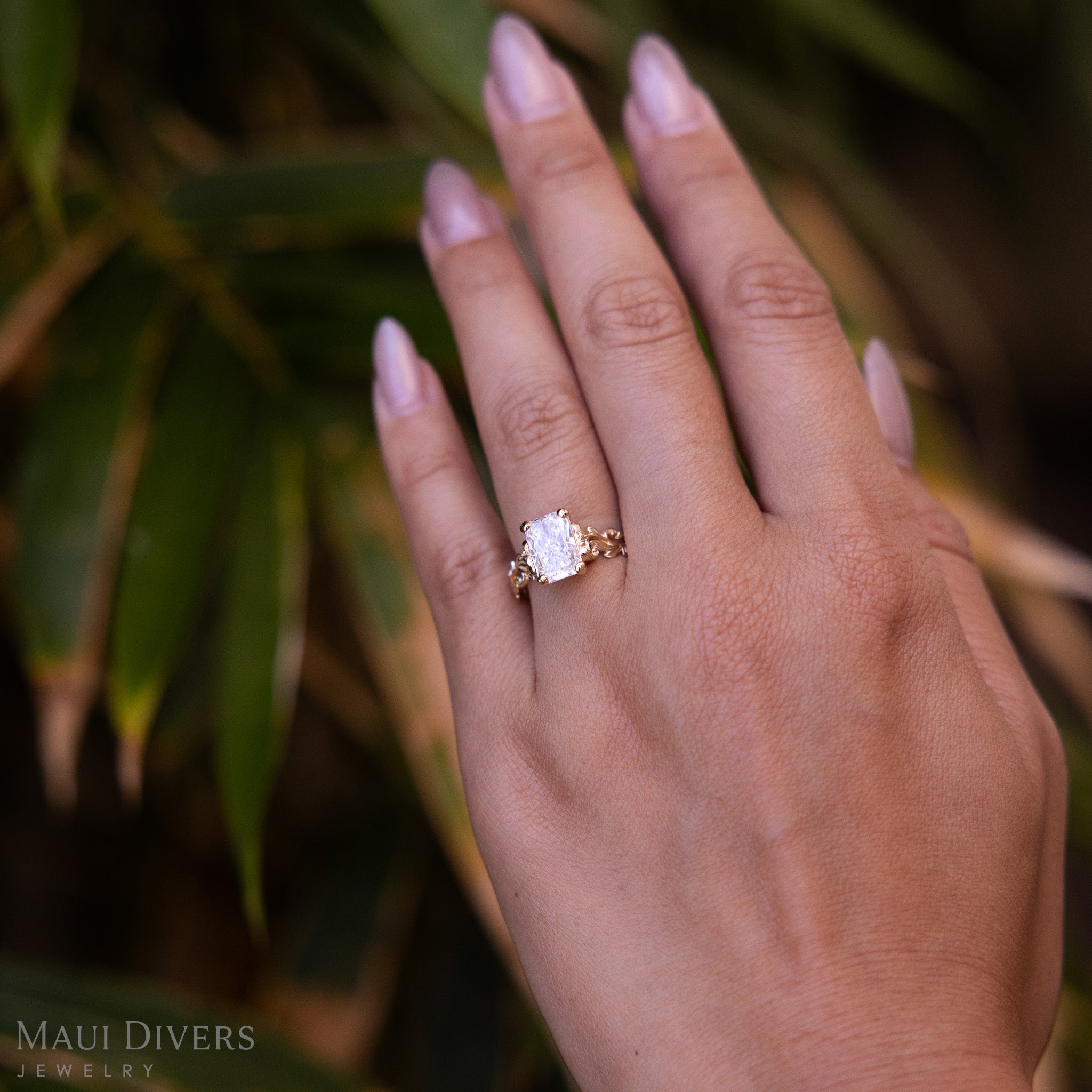 A Living Heirloom Solitaire Lab Grown Diamond Engagement Ring in 14k yellow gold worn on the ring finger of a hand, against a blurred leafy green background