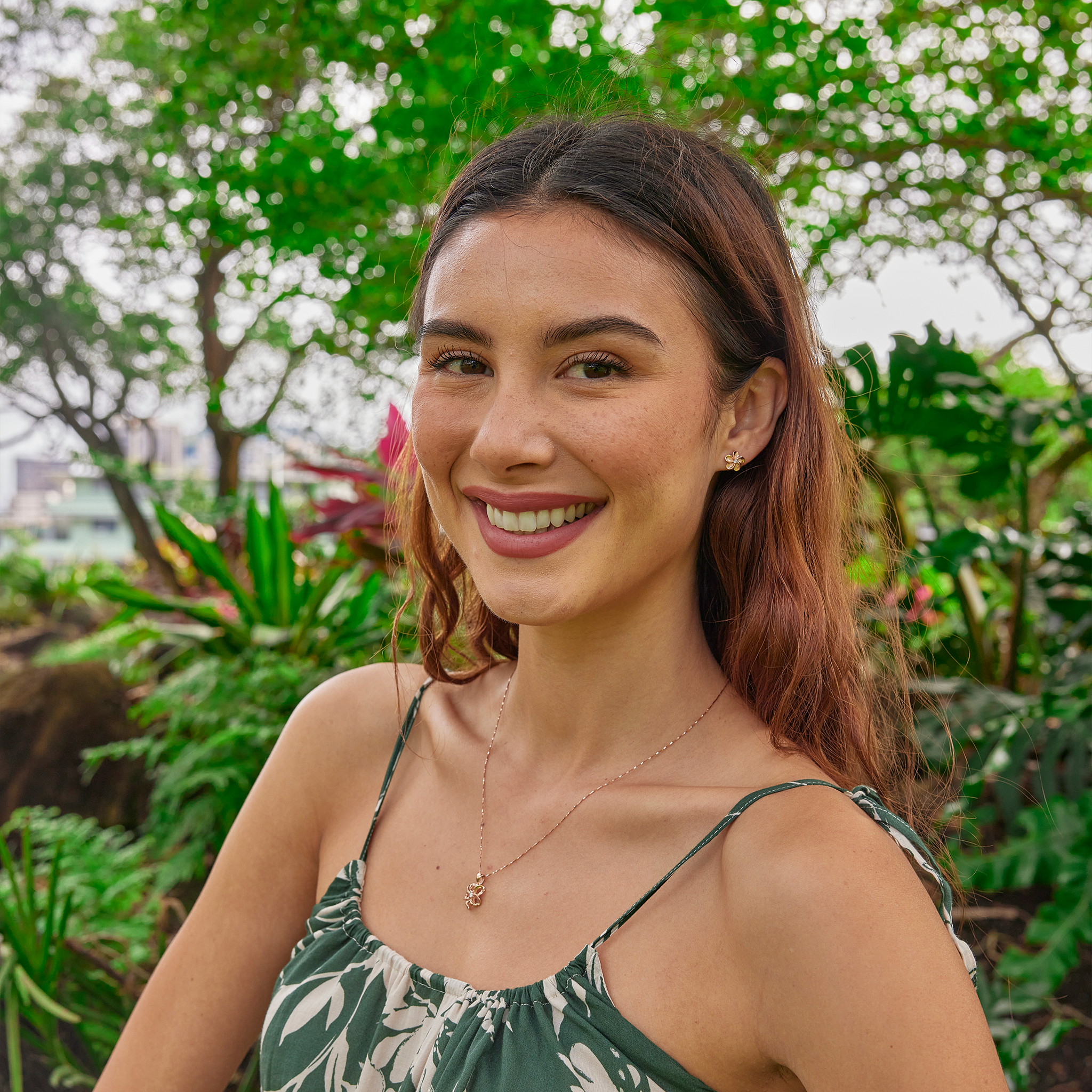 Smiling woman in lush garden wearing a cut-out design Plumeria Necklace in 14k Rose Gold