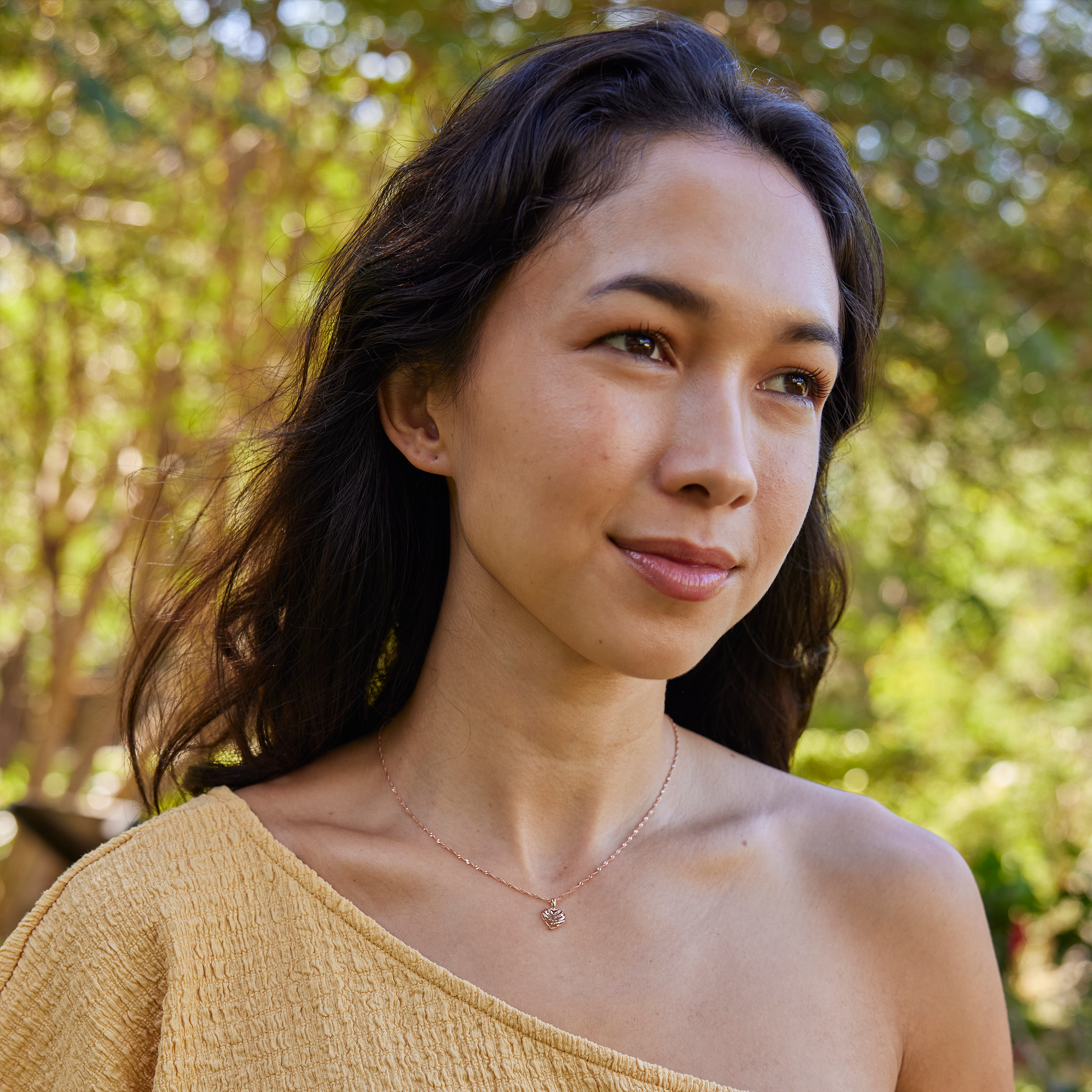 Woman wearing rose gold Aloha Heart Pendant on chain in front of green foliage