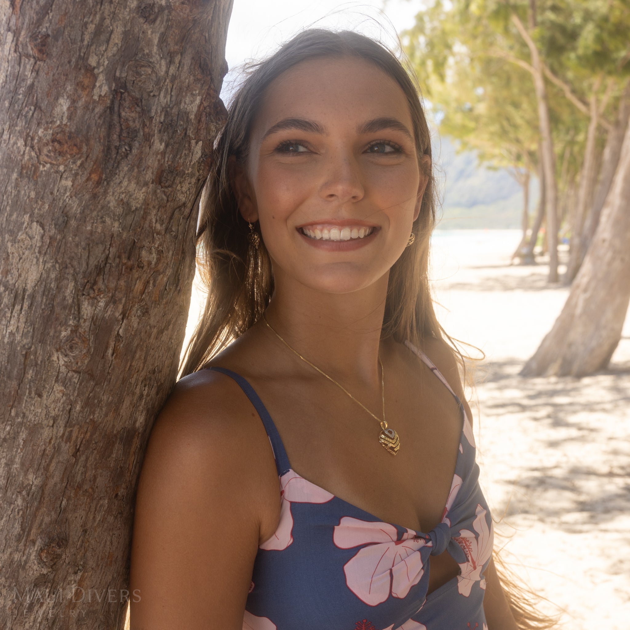 Woman leaning against a tree on a beach wearing a gold Aloha Heart Pendant