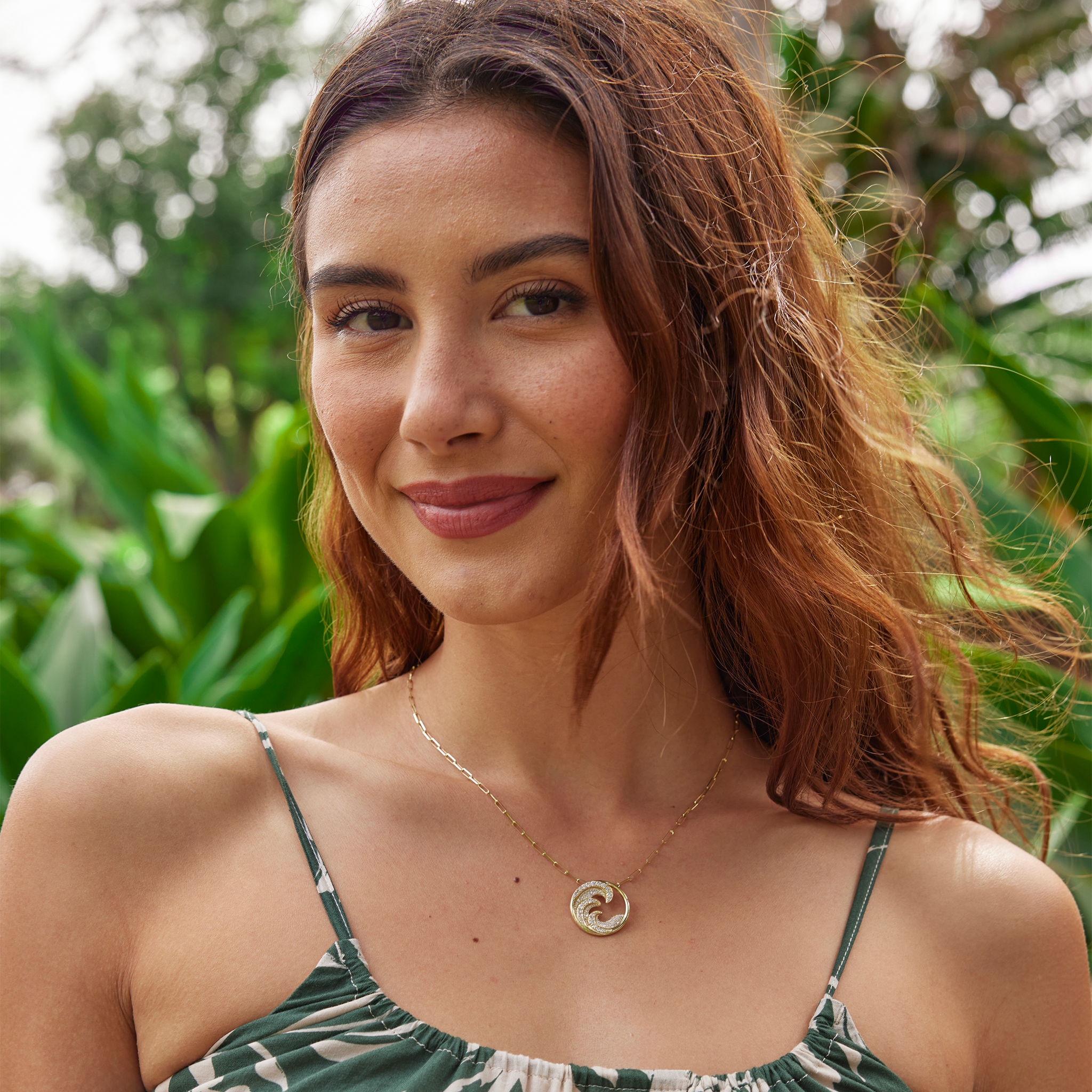 Woman in a lush garden wearing a diamond encrusted triple wave pendant in gold on paper clip chain