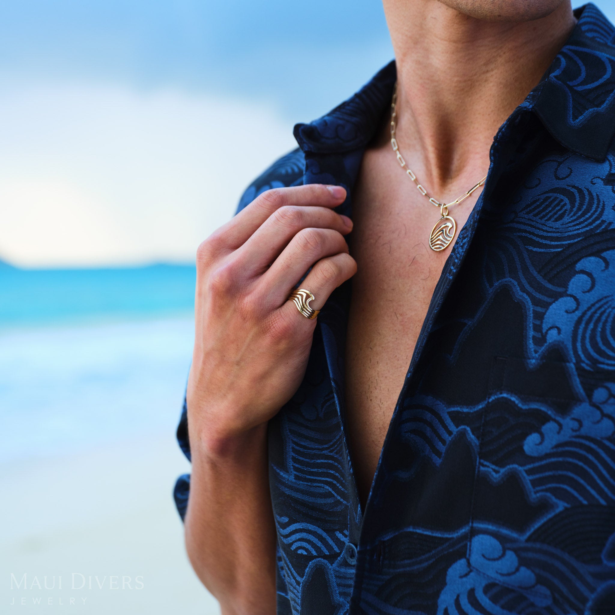 Man wearing a blue patterned shirt with an Abstract Nalu Ring and Pendant in Gold, against a blurred beach background.