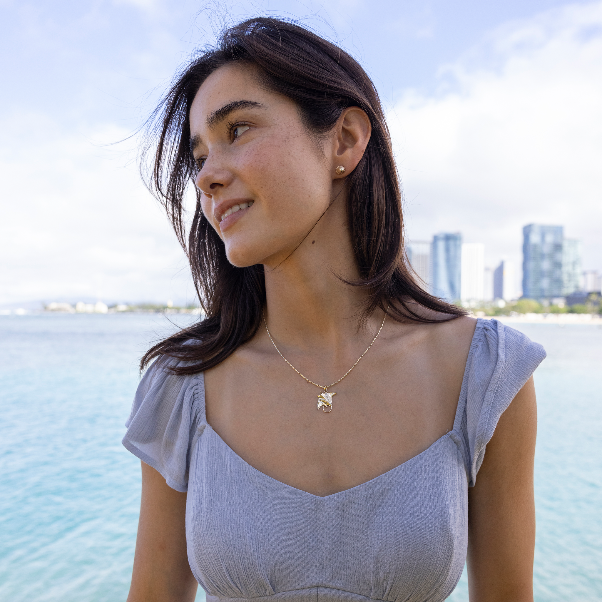 Woman wearing a blue dress and an Ocean Dance Manta Ray Pendant in gold with diamonds with ocean in the back
