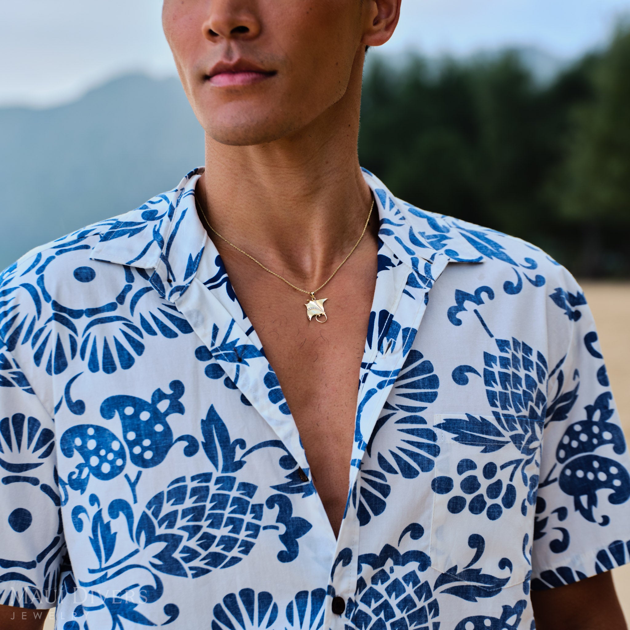 Man wearing a blue and white patterned shirt with a gold manta ray pendant on a rope chain outdoors.