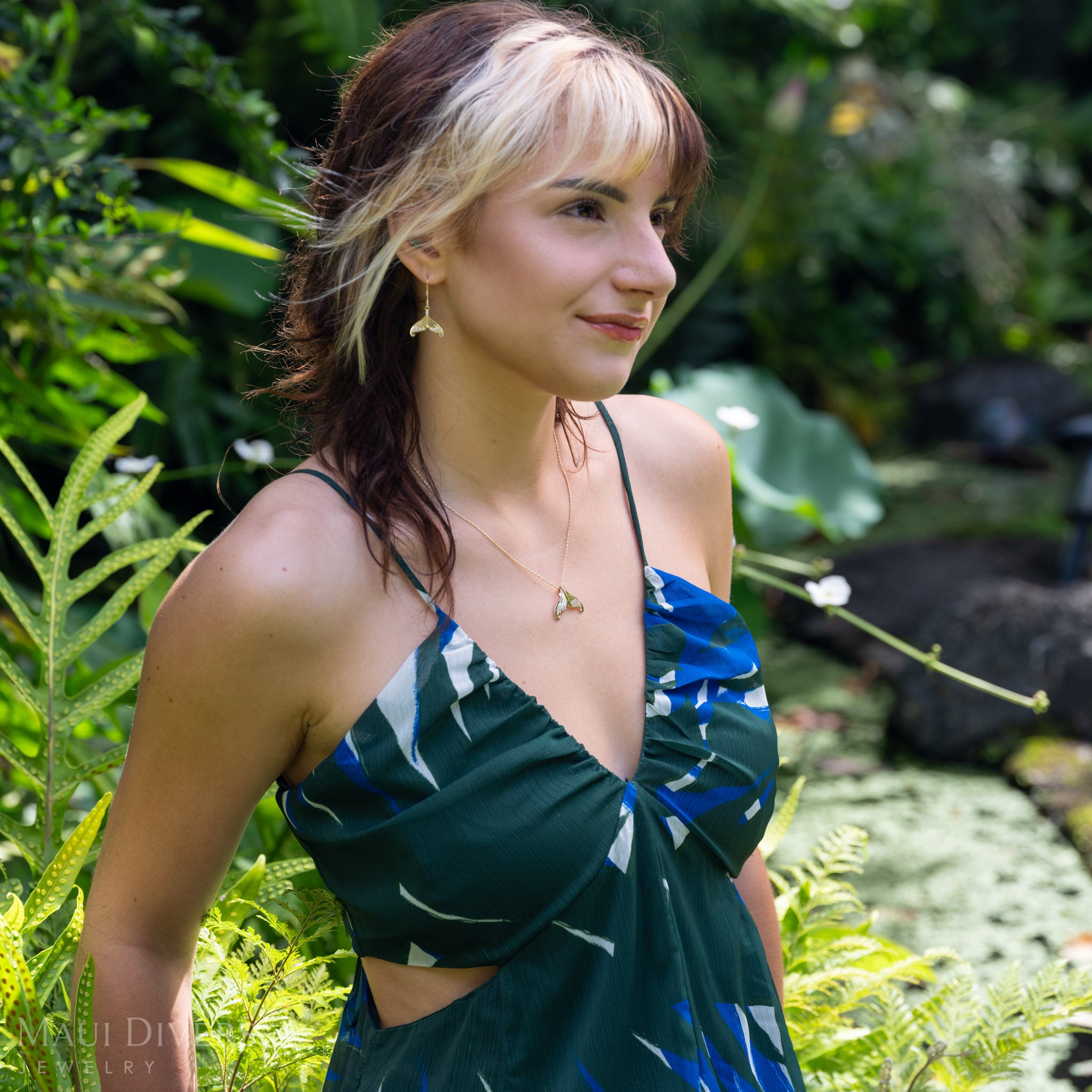A woman wearing a diamond yellow-gold mermaid-tail pendant and earring, and a dark green-and-blue patterned halter dress with a side cutout, standing in a lush green outdoor garden setting.