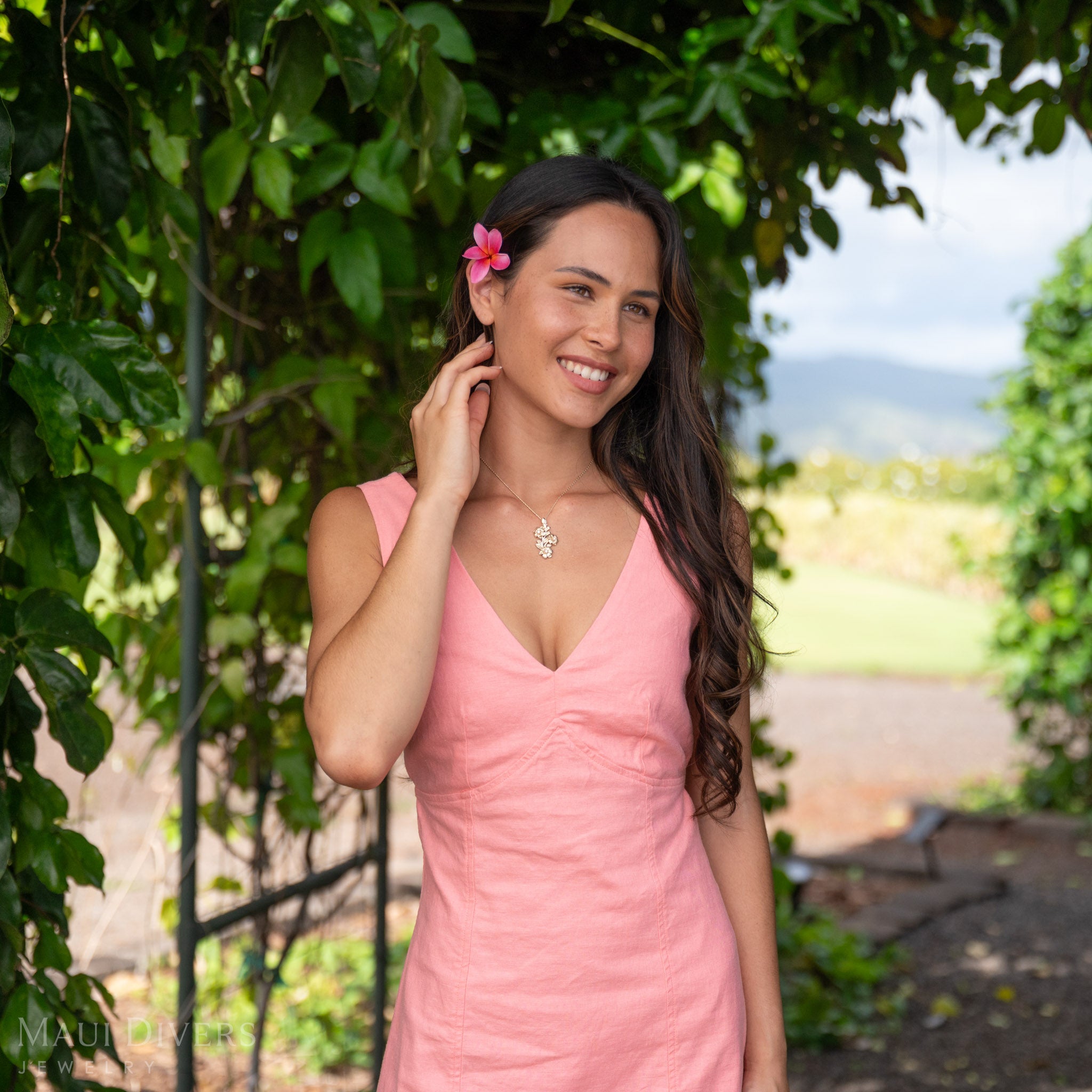 Smiling woman wearing a 14k Gold Hawaiian Gardenia pendant accented with Natural Diamonds with a pink flower in her hair against a green leaf background.