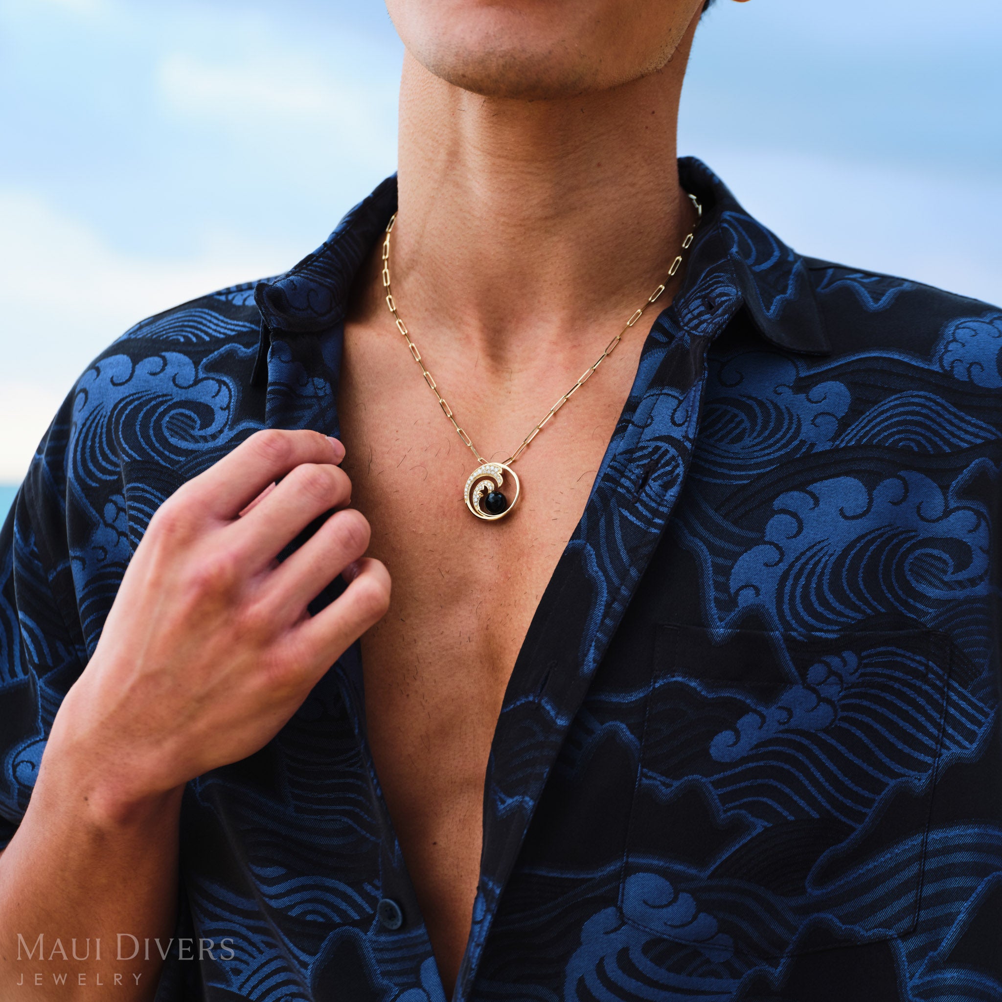 Man wearing a Nalu Black Coral Pendant in Gold with Diamonds against a blue sky