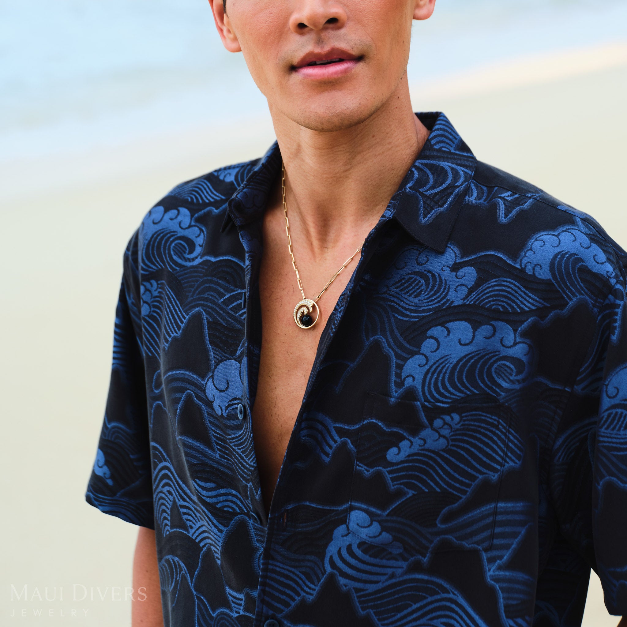 Man wearing a Nalu Black Coral Pendant in Gold with Diamonds against a blurred beach background