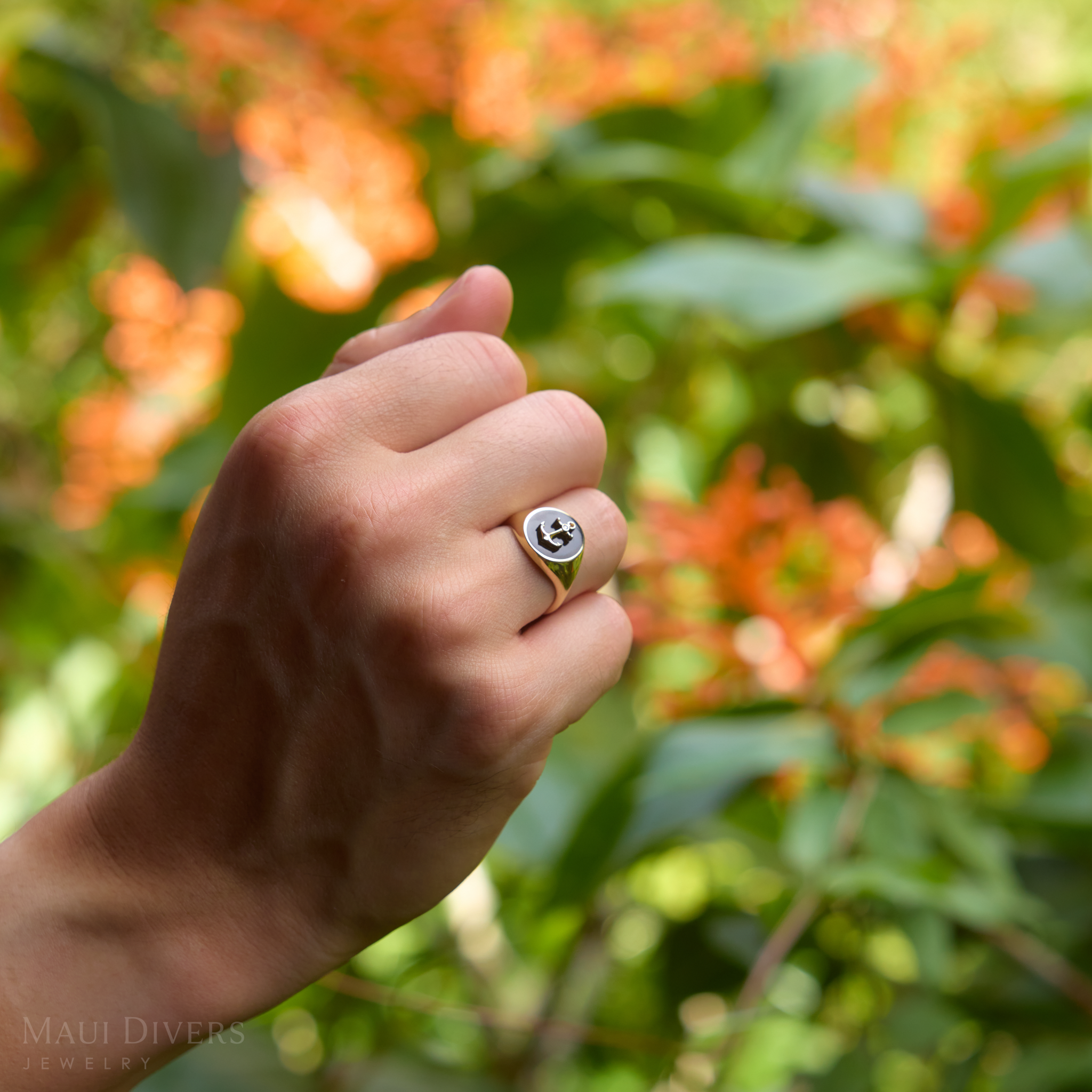 Close up of Sealife Anchor Black Coral Signet Ring in Gold with Diamond on hand amongst blurred foliage