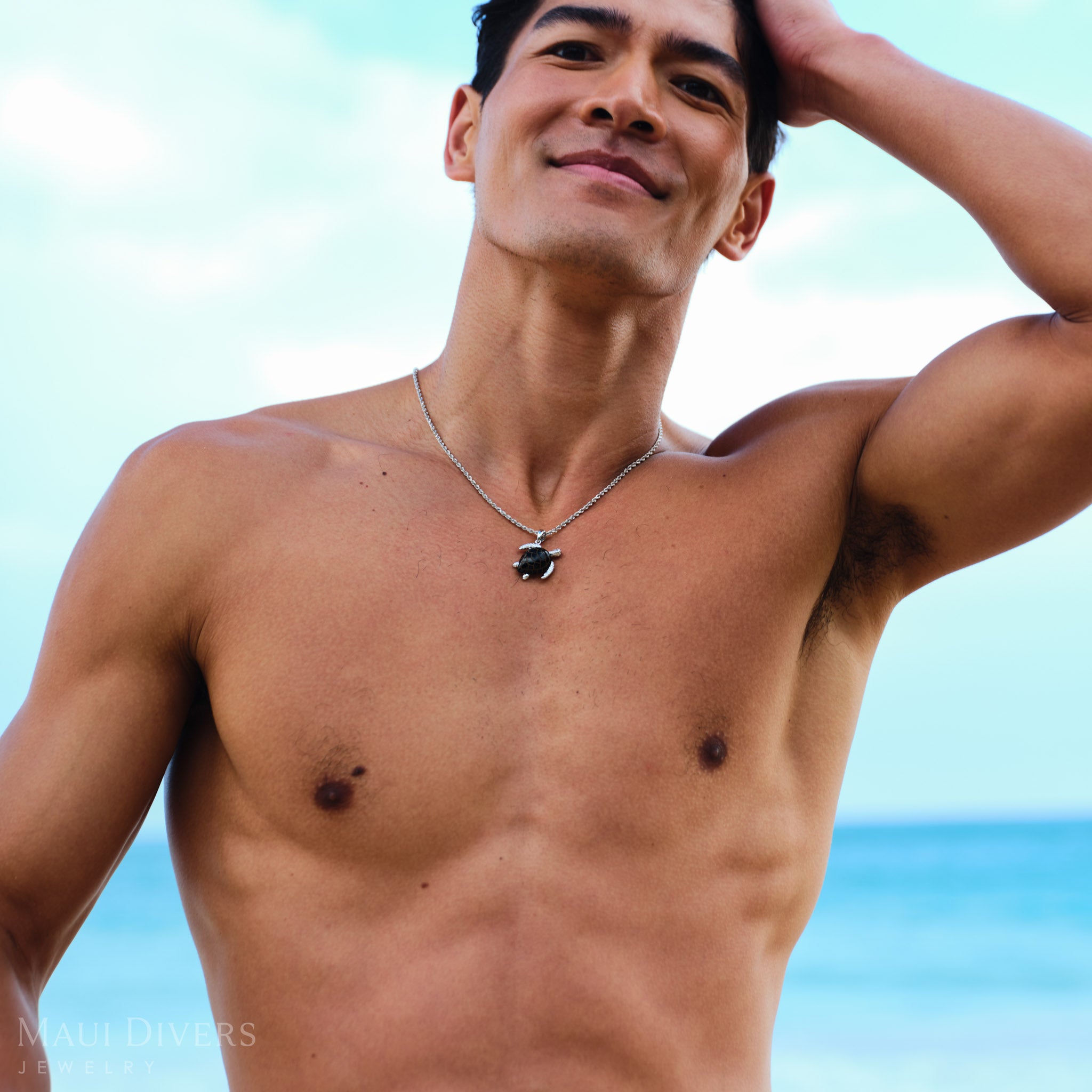 Man wearing a Honu Black Coral Pendant in White Gold with Diamonds against a blurred ocean background