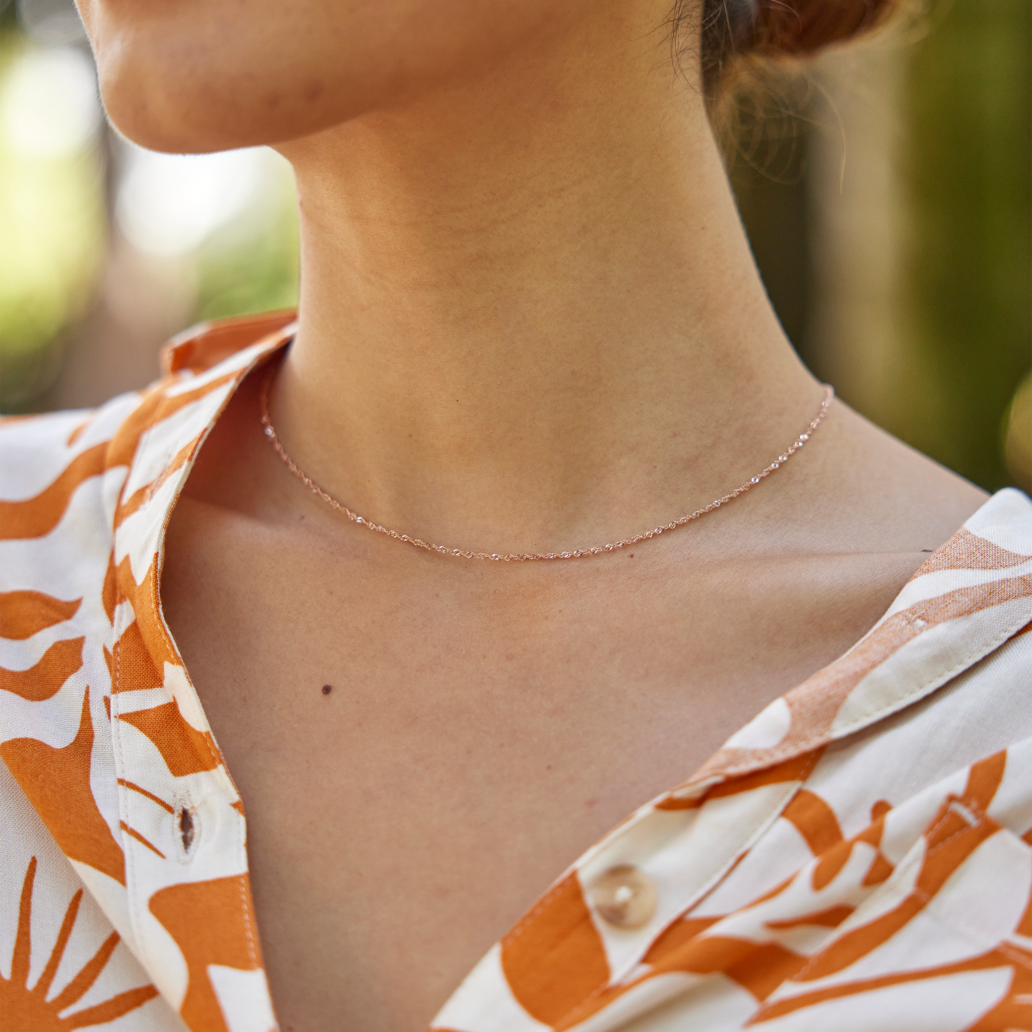 Close up of 1.0mm Singapore Chain in Rose Gold on woman's neckline with blurred greenery