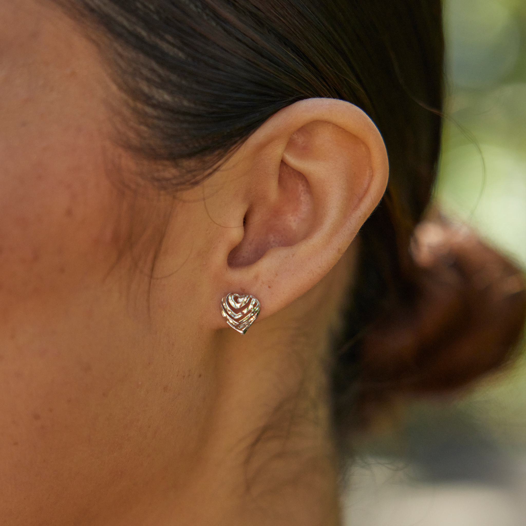 Close up of Aloha Heart Earrings in Sterling Silver on a woman's ear with blurred greenery in the back