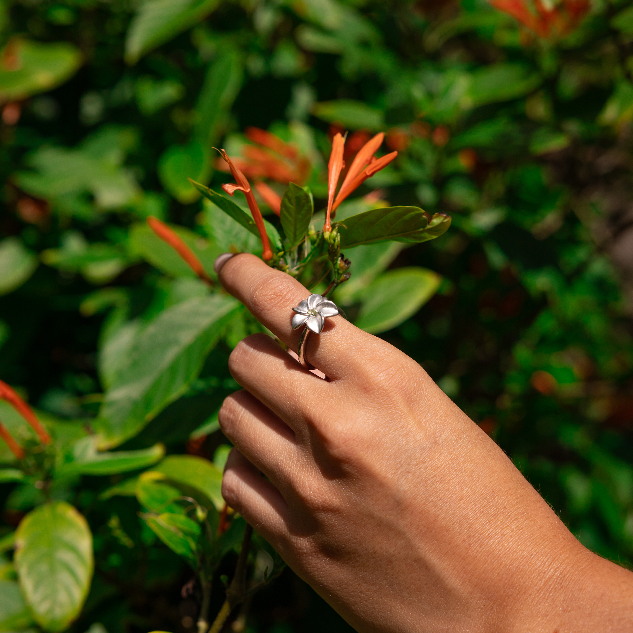 Woman's hand in a garden holding a flower and wearing a Plumeria Peridot Ring in Sterling Silver