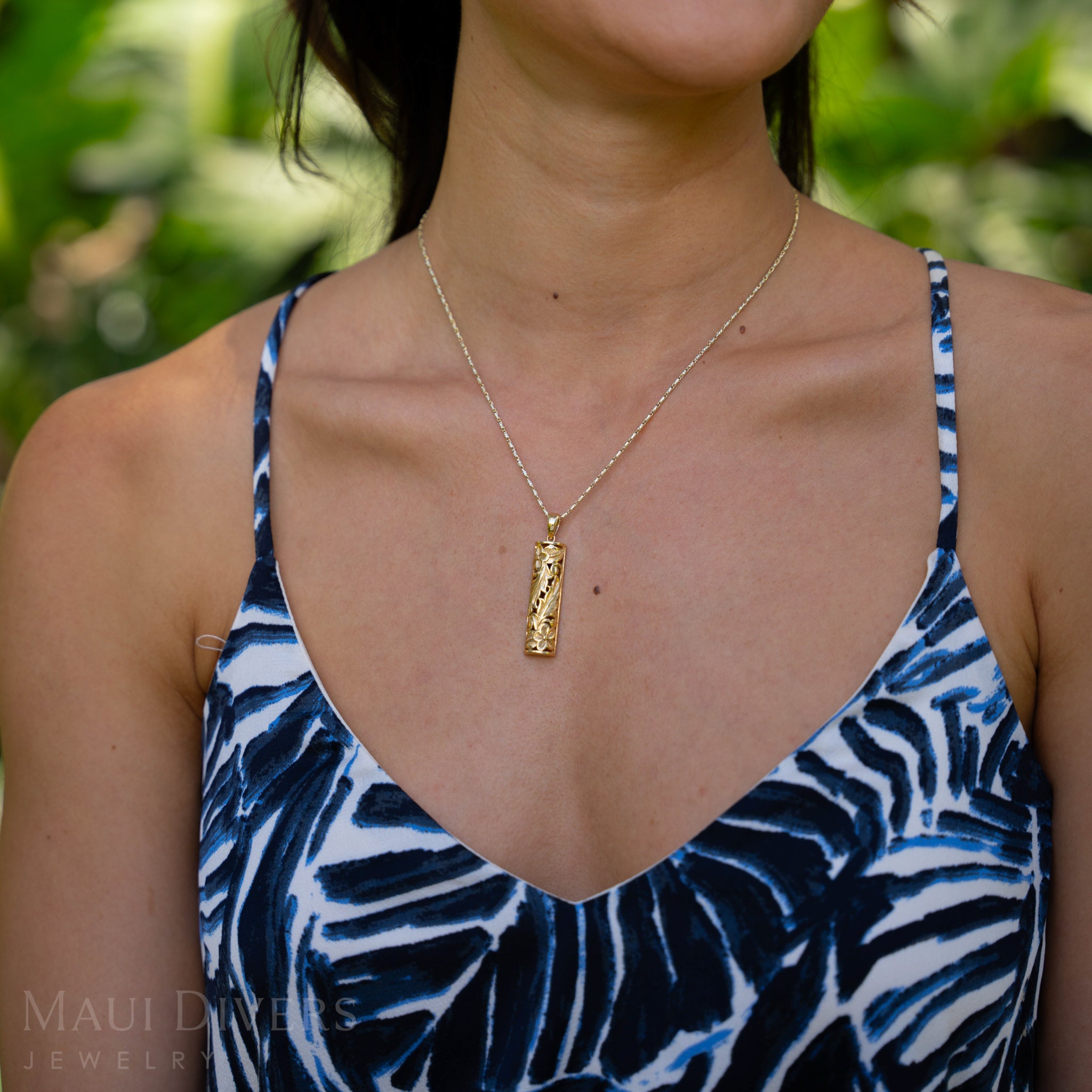 Woman wearing a Hawaiian Heirloom Plumeria Pendant in Gold against a blurred green background