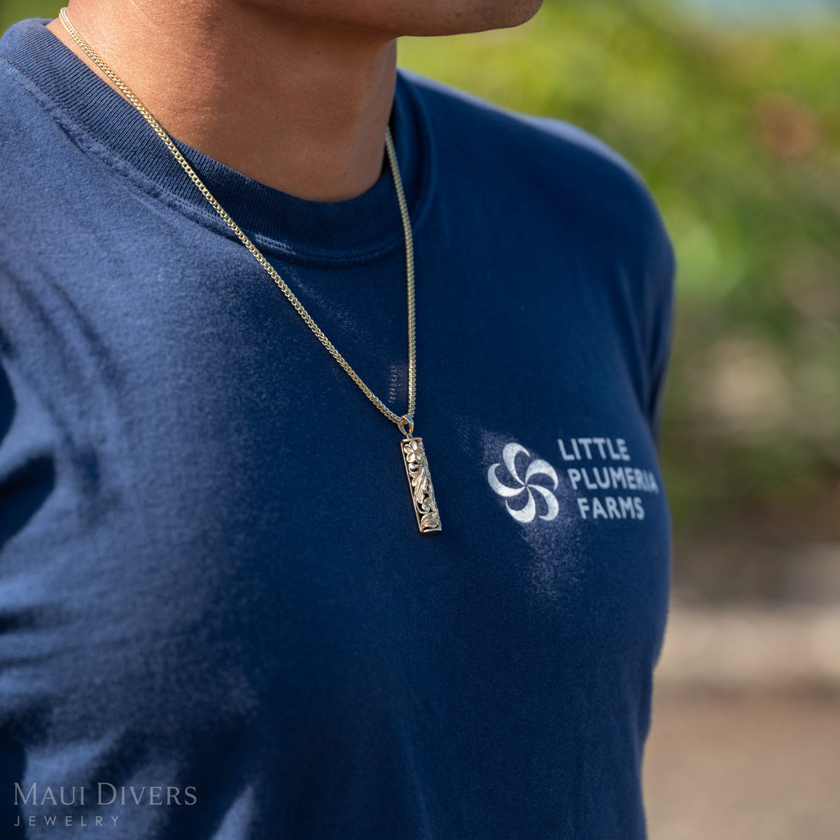 Close up shot of Dane Little wearing a gold vertical bar Hawaiian Heirloom pendant on a chain and a dark blue shirt with ʻLittle Plumeria Farmsʻ in white