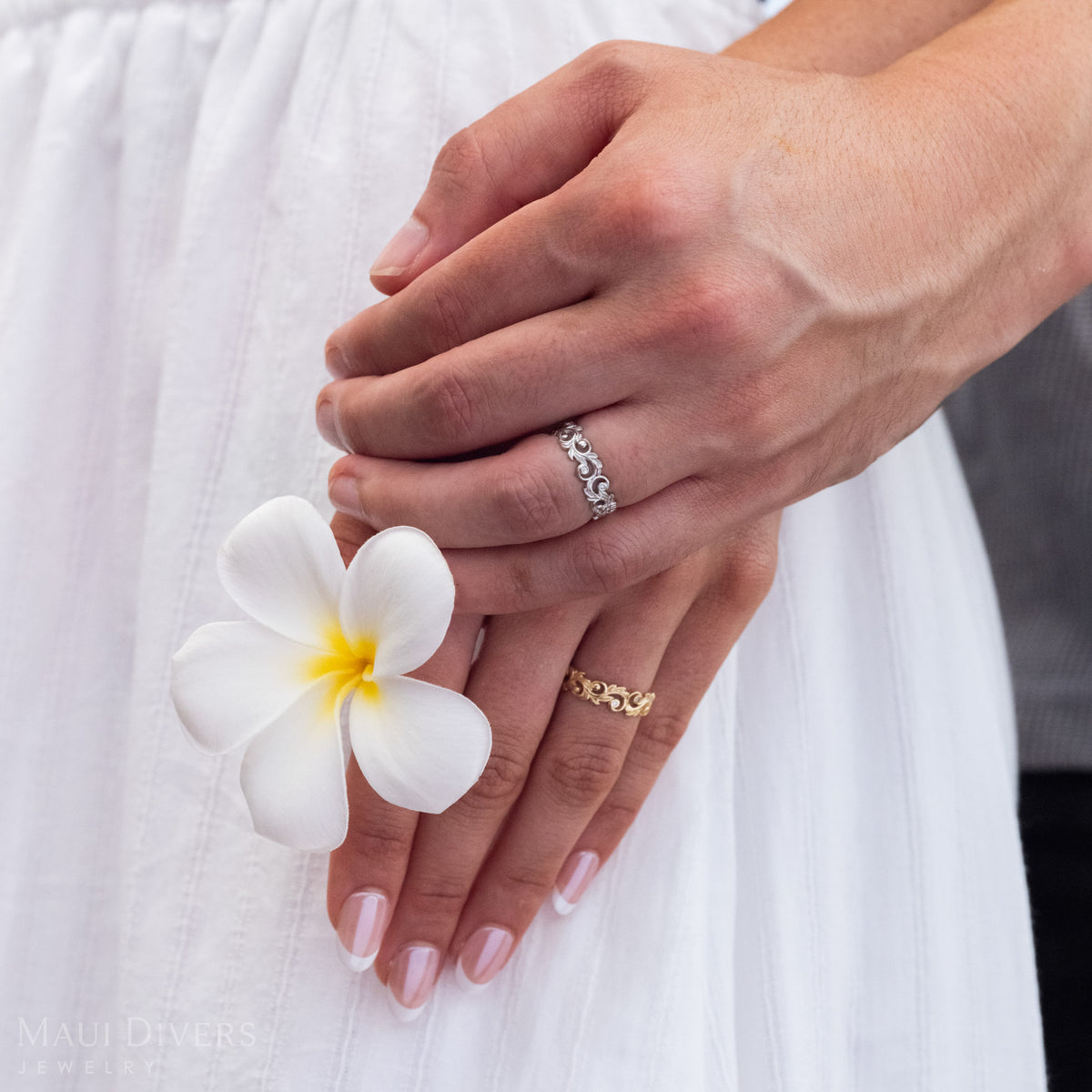 A hand holding another hand wearing Living Heirloom Ring in 14k White Gold and 14k Yellow Gold with Diamond accents, holding a plumeria flower.