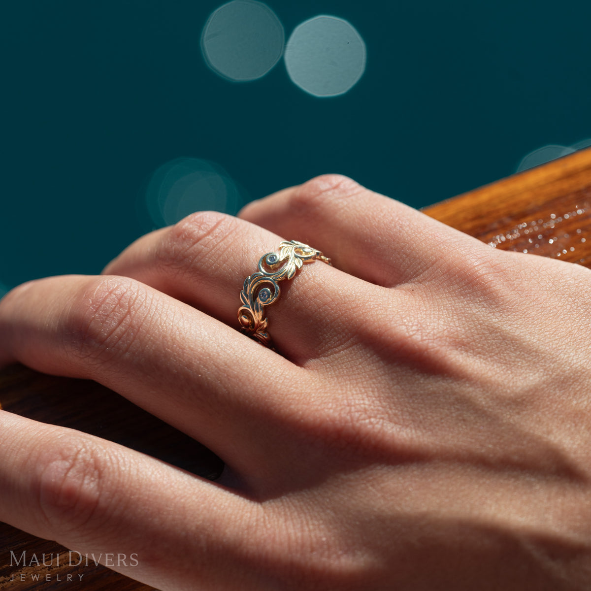 Close-up of a hand wearing a gold ring with cut-out maile leaf and fern scrollwork with diamonds, against a sparkling deep ocean background