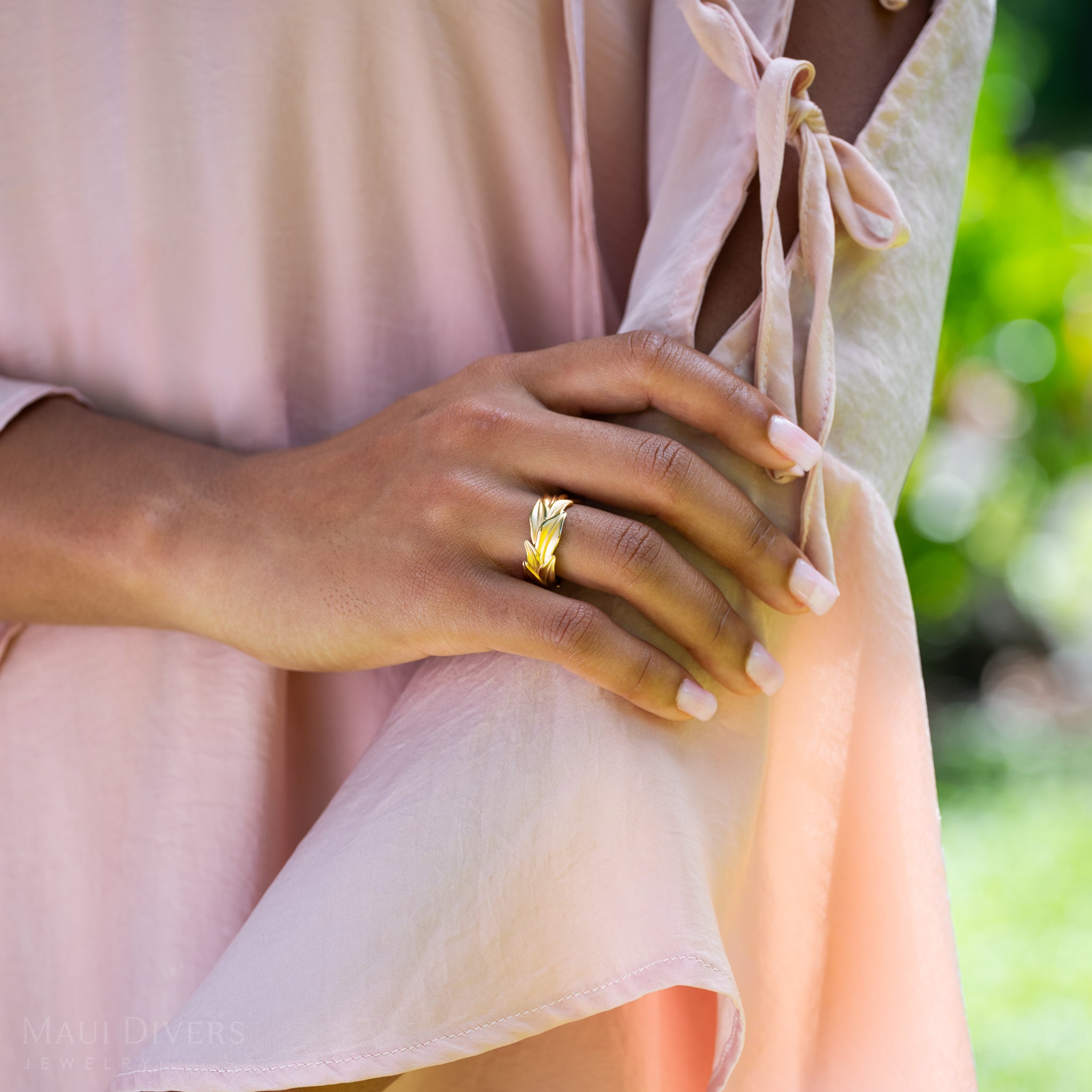 Close-up of a hand wearing a 10mm Maile Ring in Gold with a blurred green background